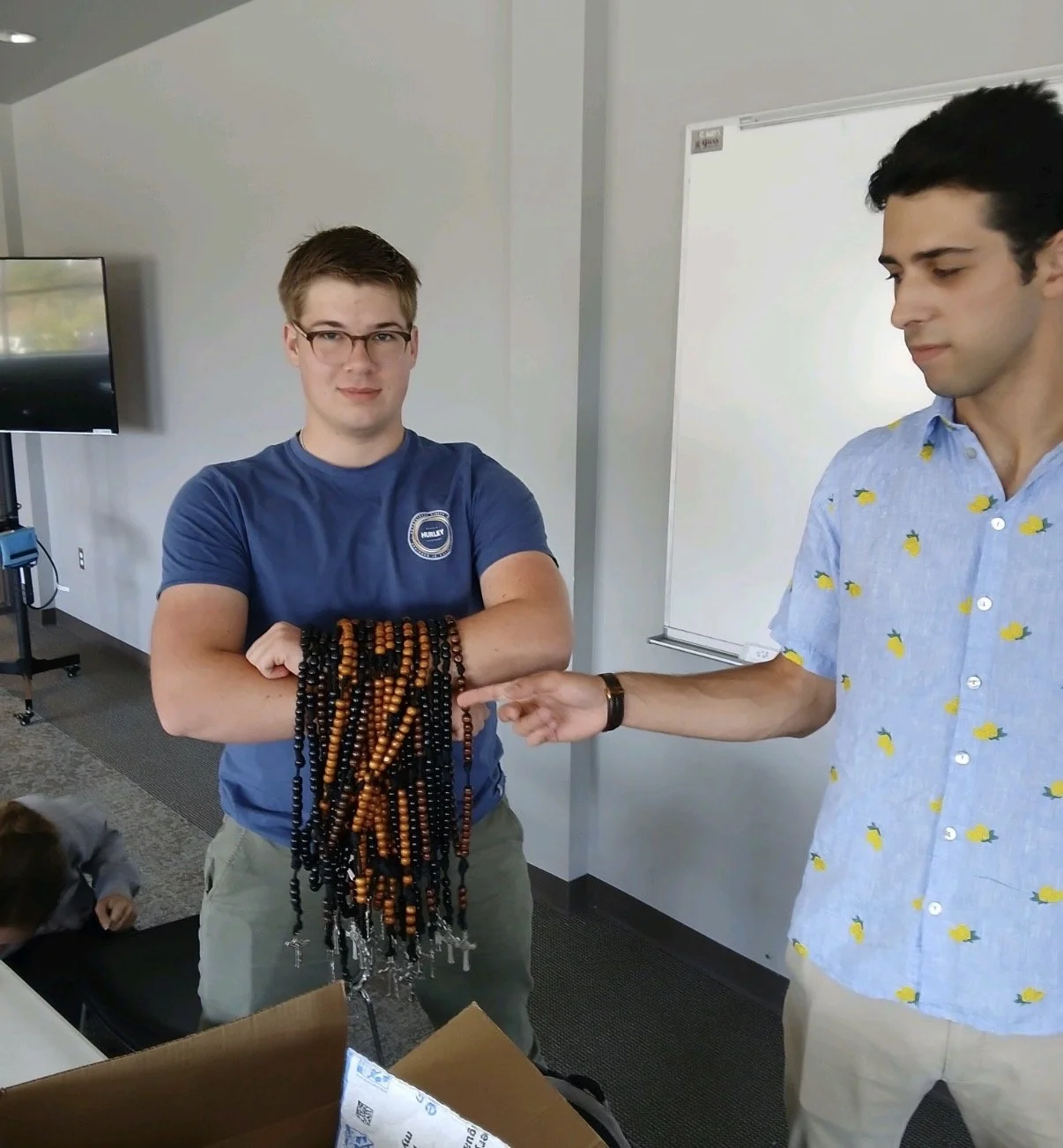 A young man in a blue t-shirt with glasses holds a large bundle of black and amber prayer beads. Another young man in a light blue shirt with lemon prints points at the beads, in a room with a whiteboard and a TV in the background.