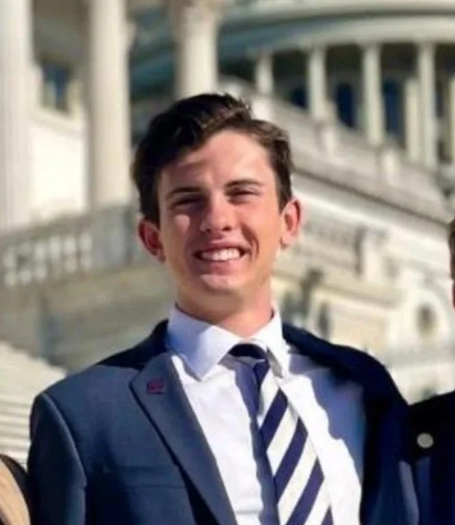 A young woman with shoulder-length black hair smiling in a greenhouse with green plants.