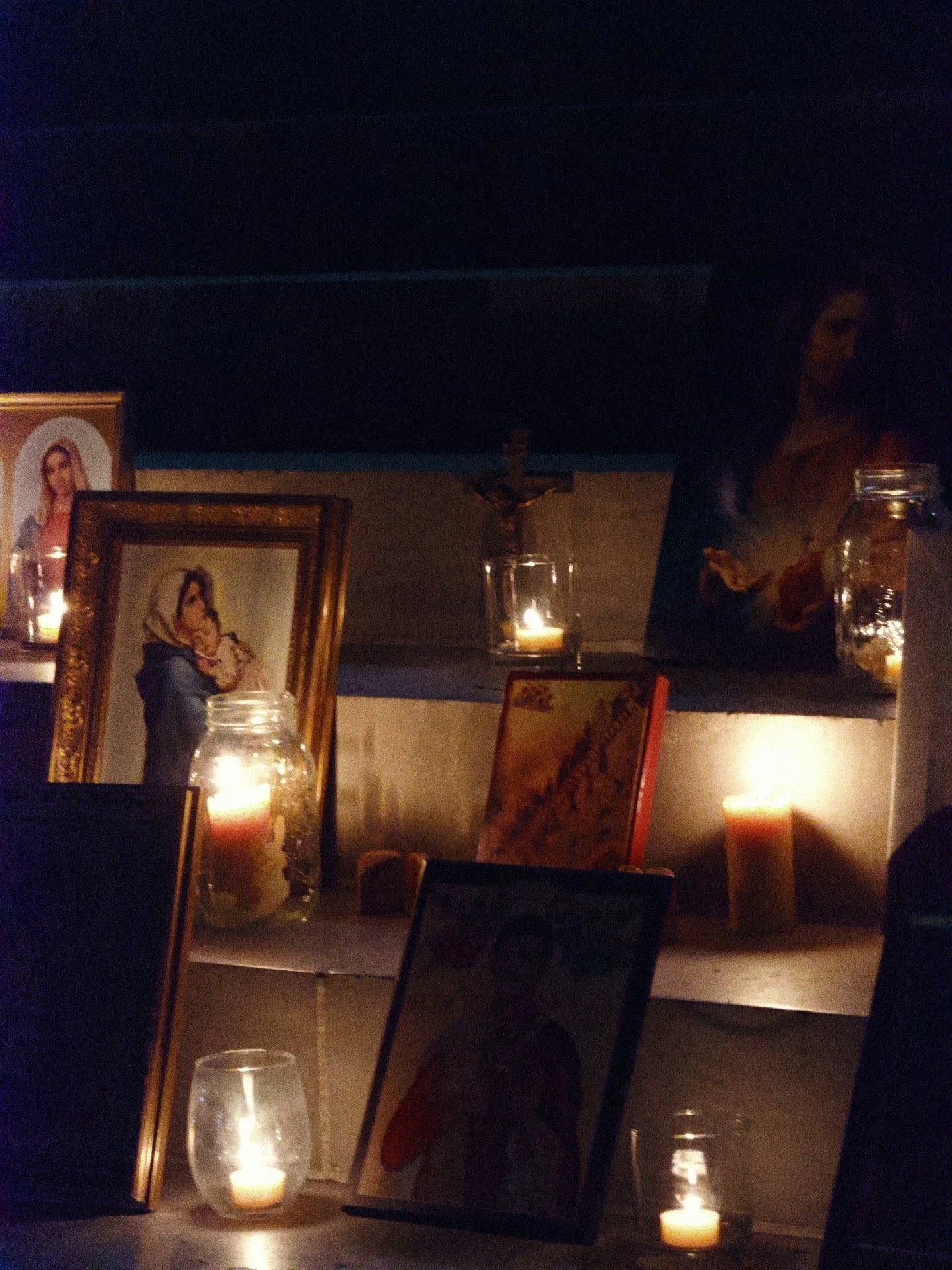 Religious shrine with candles, framed images of the Virgin Mary, and a crucifix on a wall.