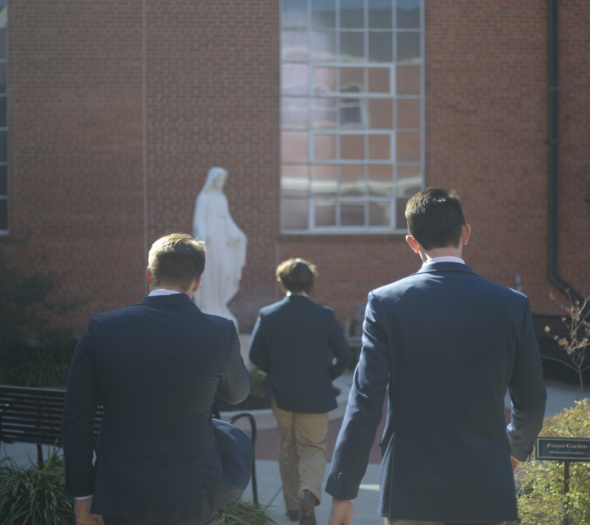 Three men in suits walking towards a religious statue outside a brick building.