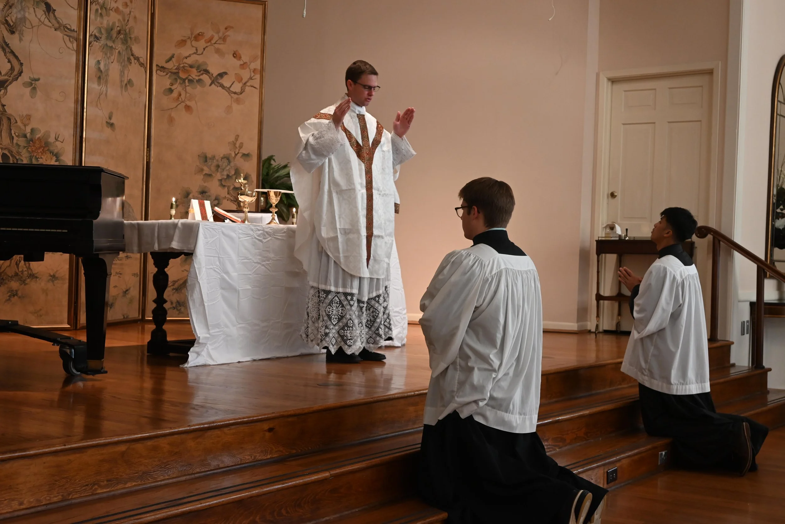 A priest conducting a religious ceremony with two altar servers kneeling on the wooden floor in front of him, inside a church with an altar, candles, and artwork in the background.