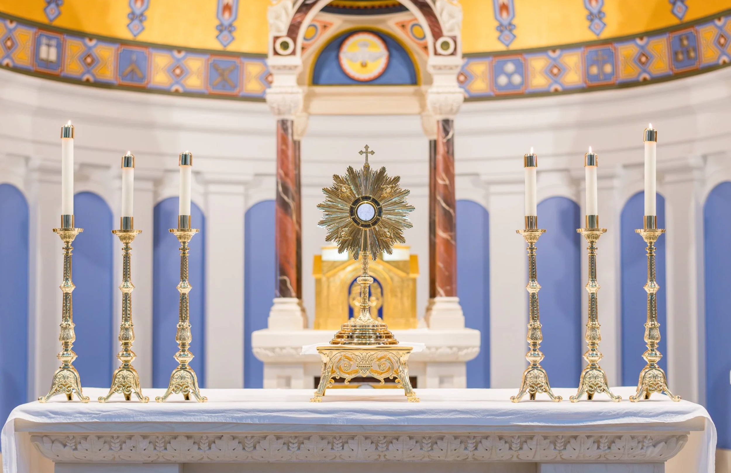 Gold monstrance on altar with six tall candles in ornate gold holders, church interior with colorful decorations and arches in the background.