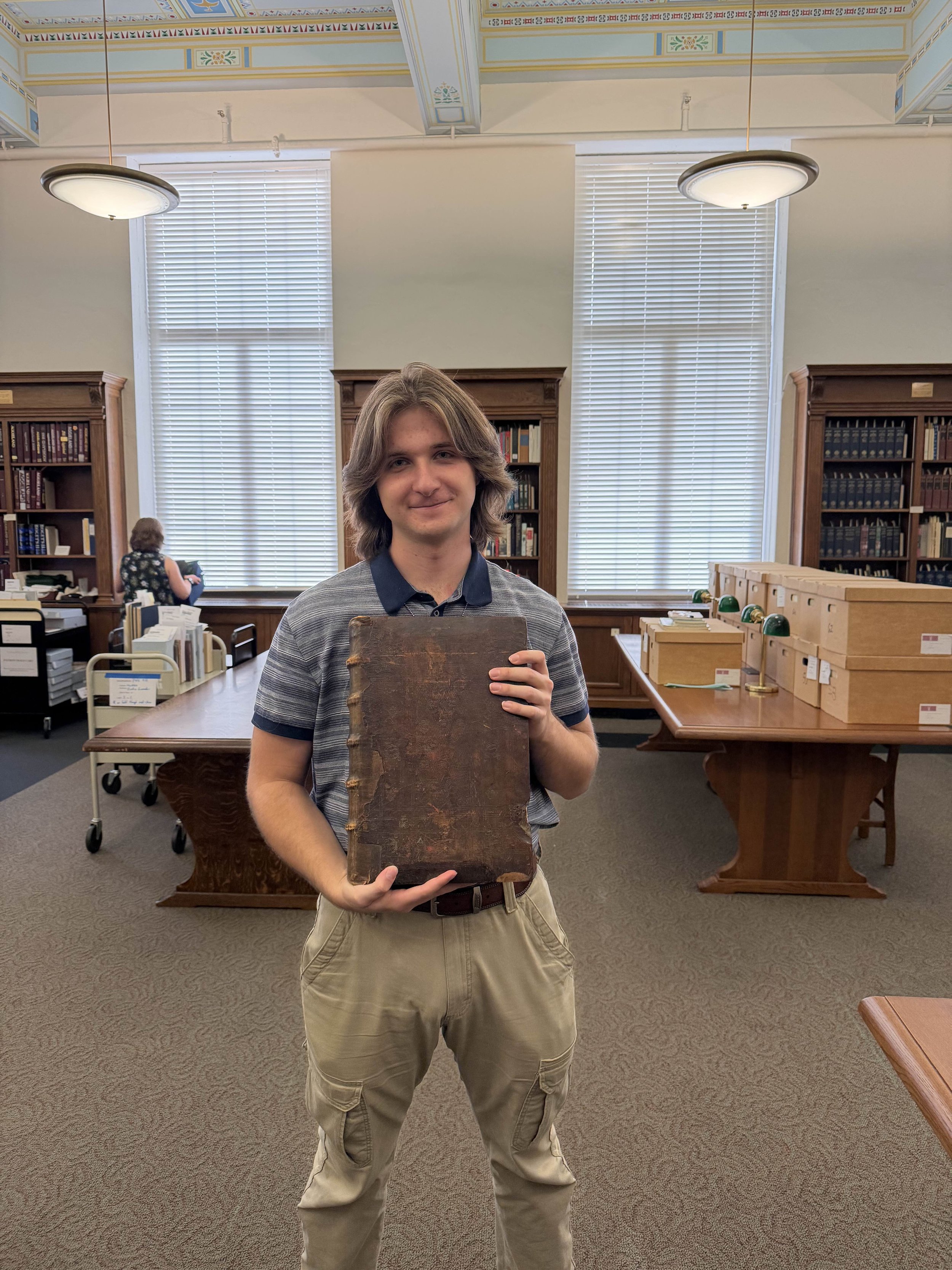 A young man standing in a library holding an old, large, leather-bound book in front of him. Behind him are tall wooden bookshelves, large windows with blinds, and a person browsing books in the background.