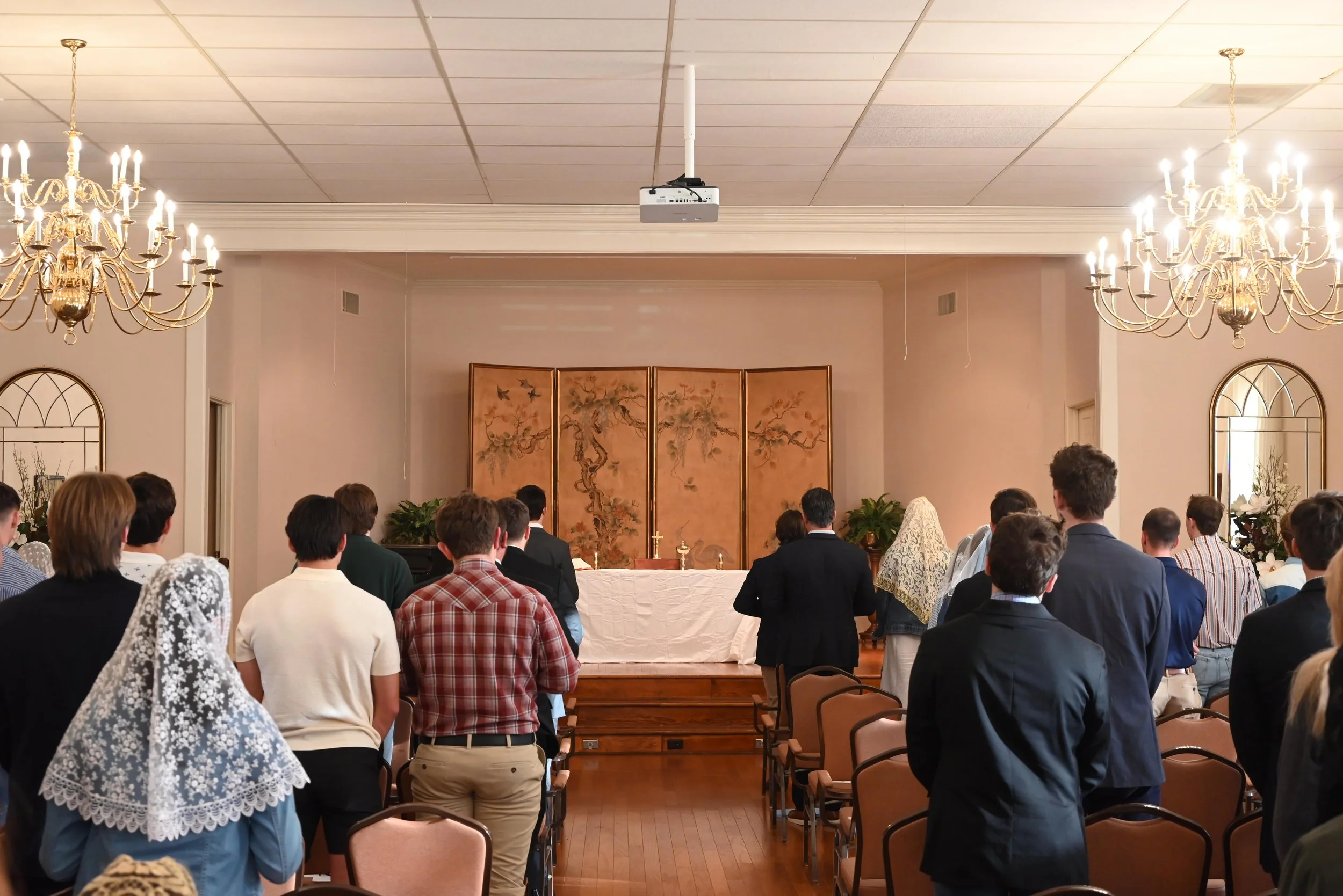People standing in a church or chapel during a religious service, facing the altar with a gold divider with tree and bird artwork, chandeliers hanging from the ceiling, and mirrors on the side walls.