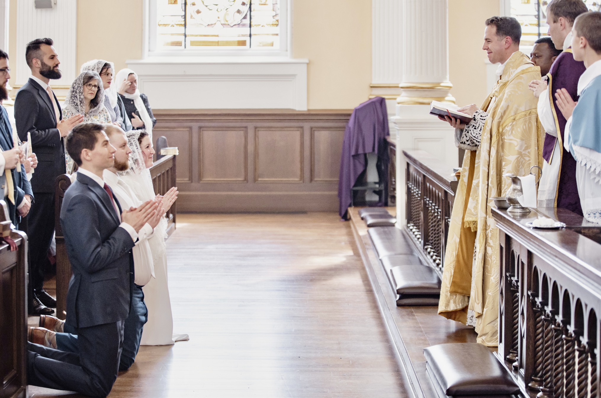 A wedding ceremony with the bride and groom kneeling and praying before the priest, with guests standing and praying in the church.