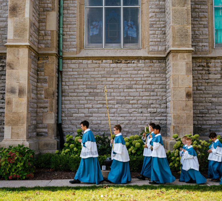 Children dressed in blue and white robes walking in procession outside a stone church, some carrying candles.