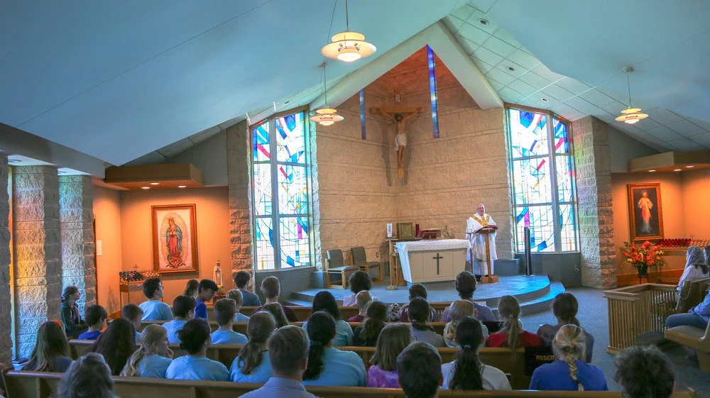 Inside a church with a congregation seated in pews, a priest at the altar, crucifix above, stained glass windows, and religious images on the walls.