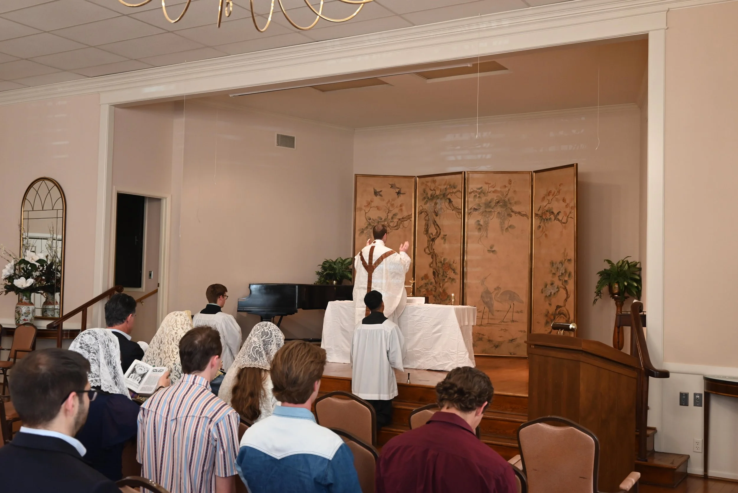 A priest leading a religious service at an indoor chapel altar with a gold folding screen, surrounded by worshippers, some with lace covers on their heads.