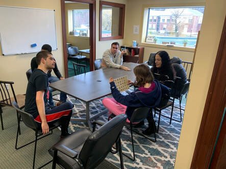 Five people sitting around a conference table in a classroom or meeting room, with a whiteboard and large windows showing a pool outside.