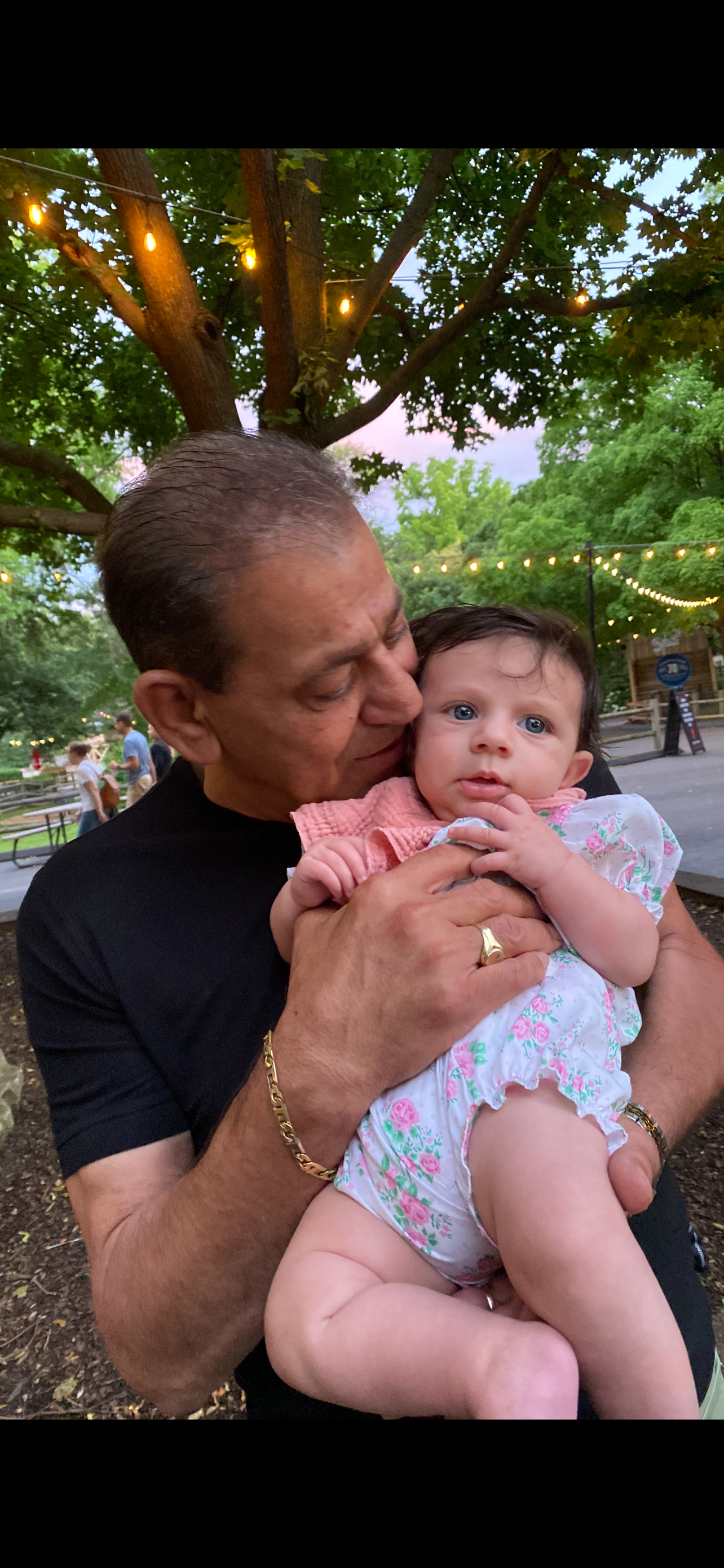Nicolas Maamari holding his grandson Vinny outdoors with trees and string lights in the background.