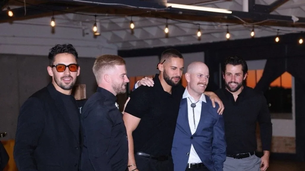 Five men in semi-formal attire standing together at an indoor party, with string lights hanging from the ceiling in the background.