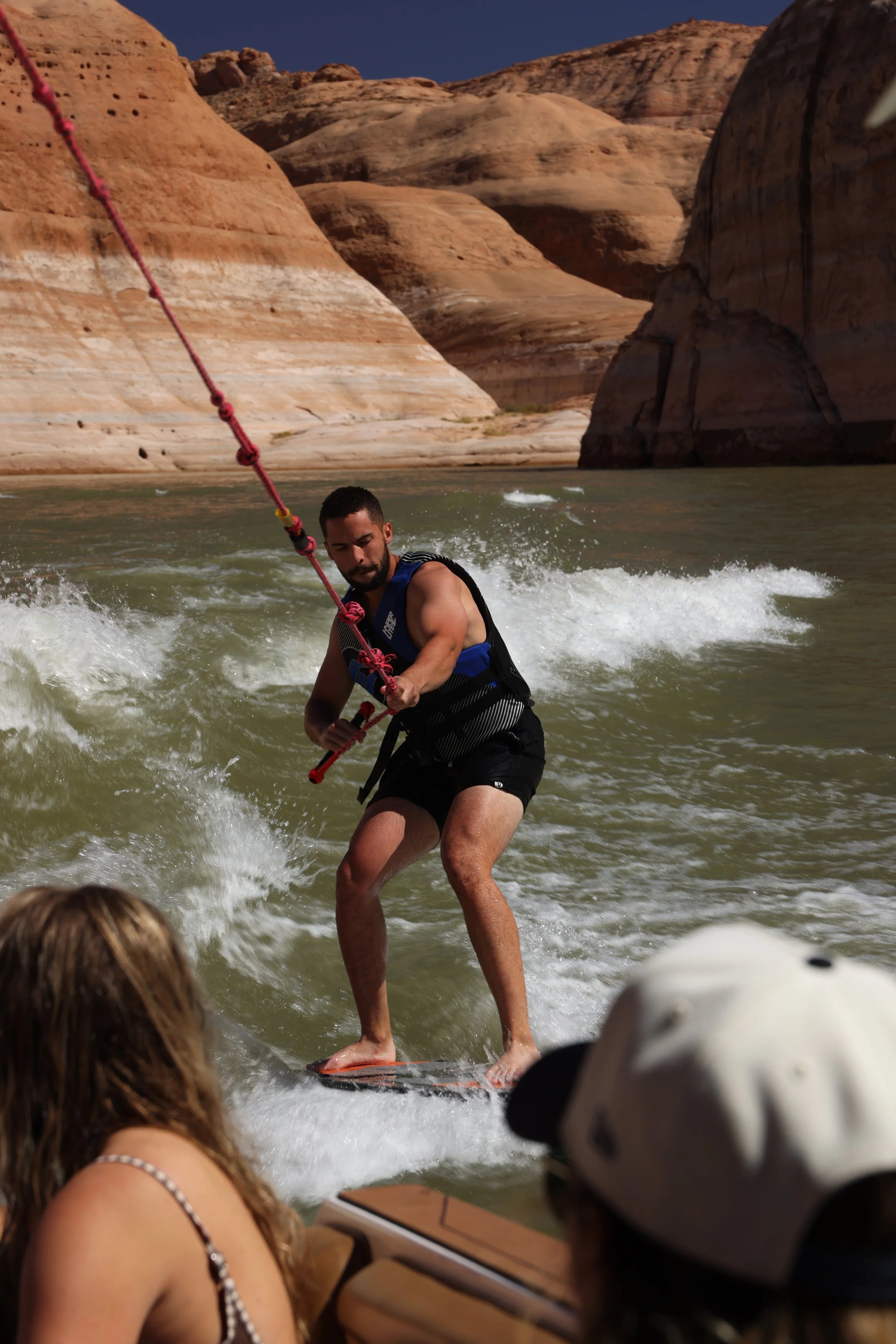 A man standing on a wakeboard on a river, holding a tow rope, with desert canyon rocks in the background and spectators watching.