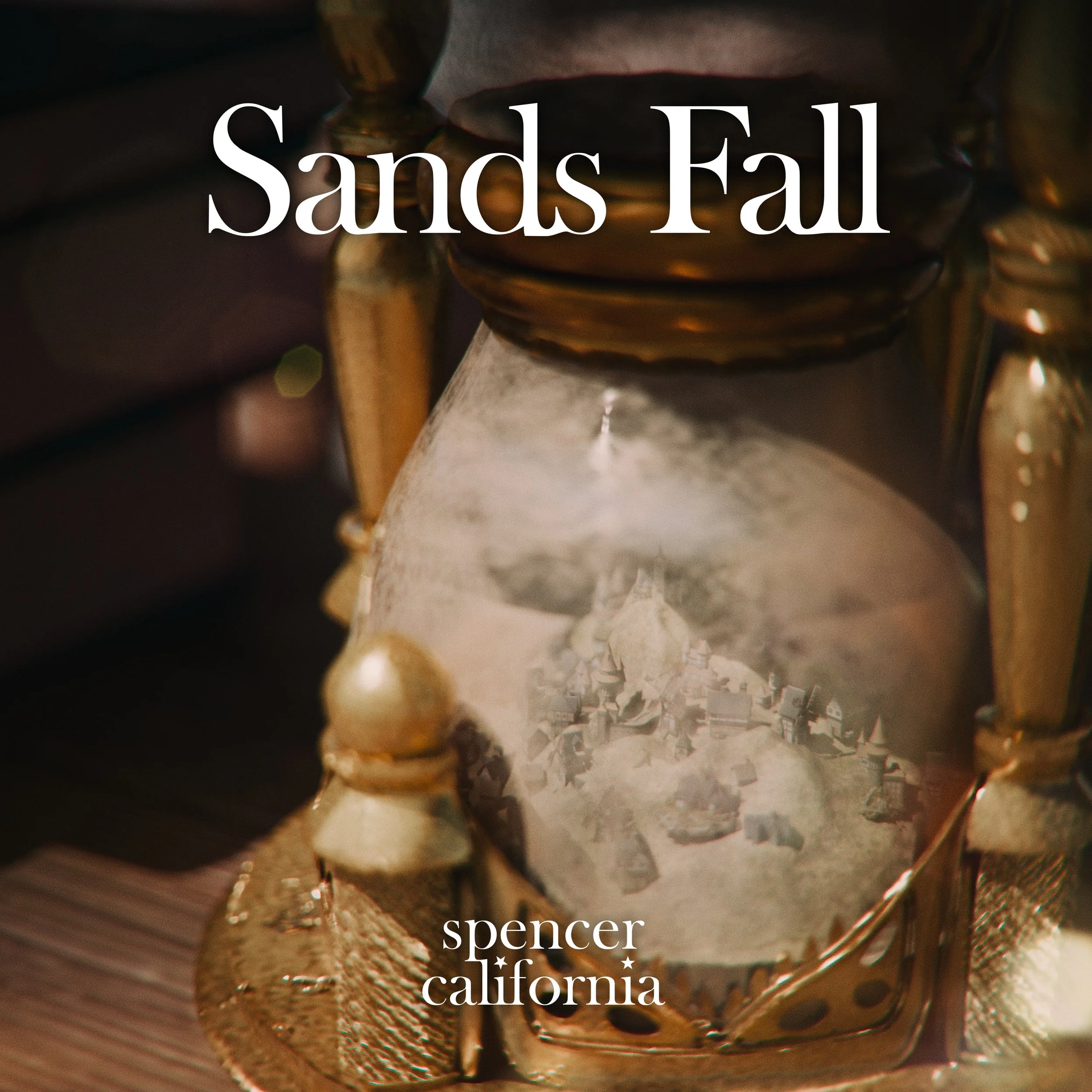 An hourglass with golden accents shows sand falling through, with a reflection of a small town or village inside the glass. The magazine cover reads 'Sands Fall' and mentions Spencer, California.