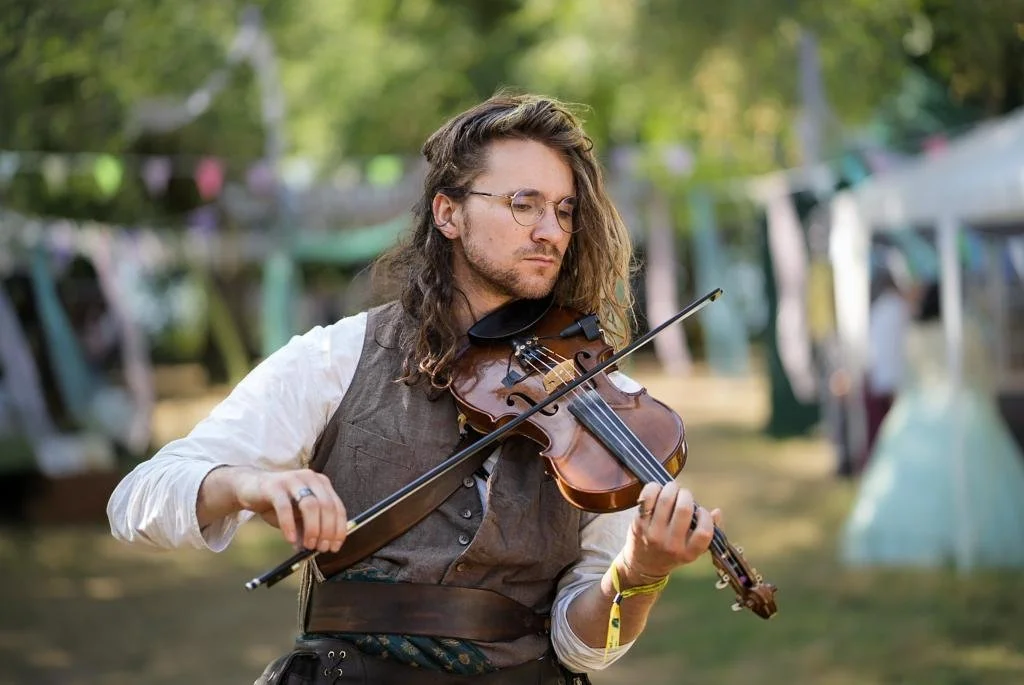 A man with long hair and glasses playing a violin outdoors at a festival or fair, with tents and trees in the background.