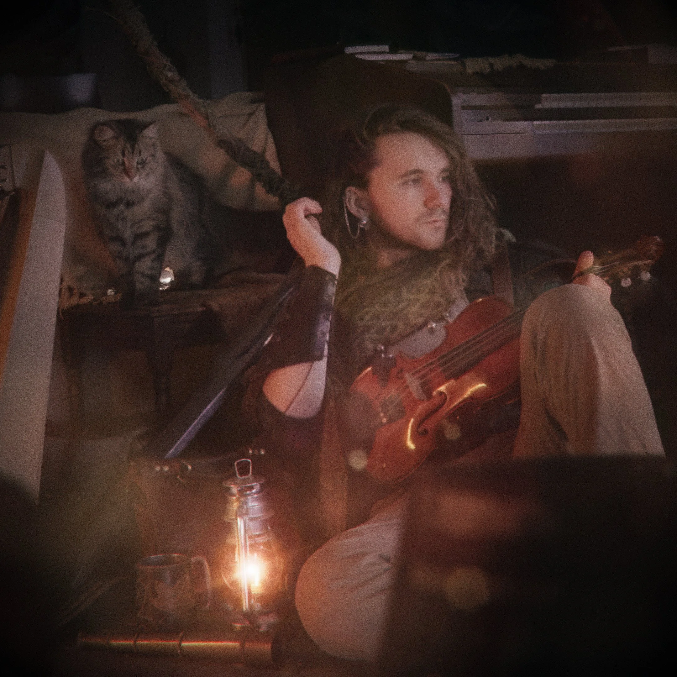 A person with long curly hair sitting outdoors at night playing a violin, with a cat sitting on a nearby chair.