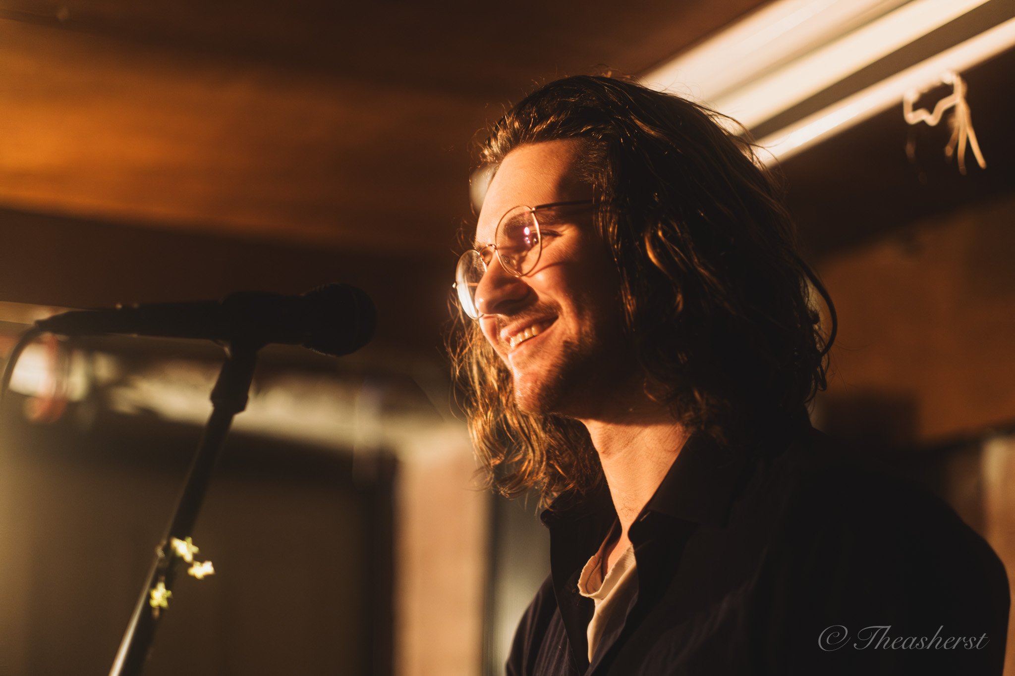 A man with long wavy hair, wearing glasses, smiling and standing near a microphone in a warmly lit indoor setting.
