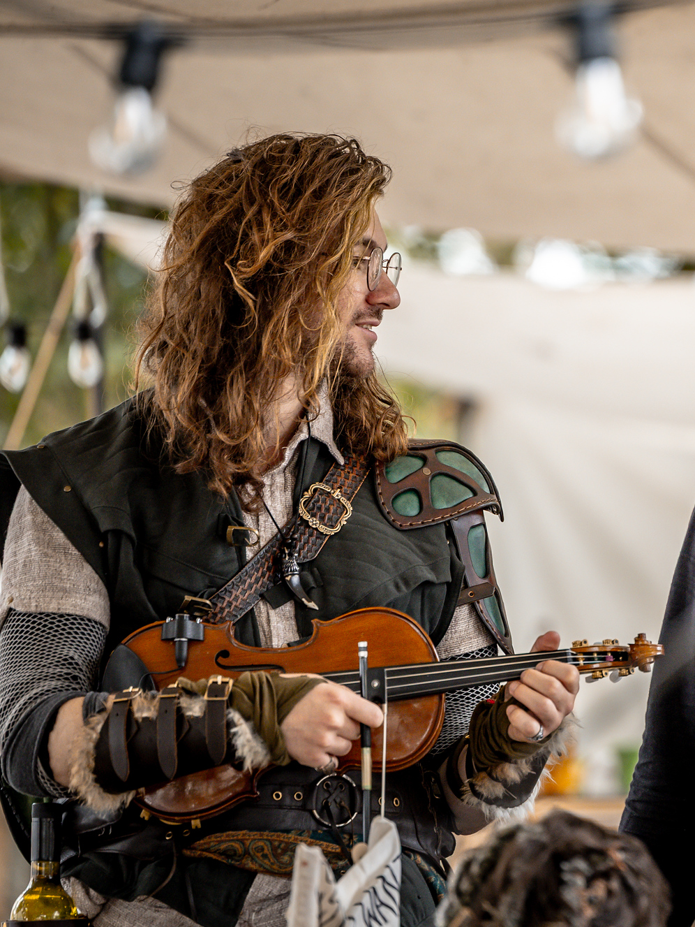A man with long curly hair and glasses playing a small string instrument, possibly a violin or viola, in a festival or outdoor setting.