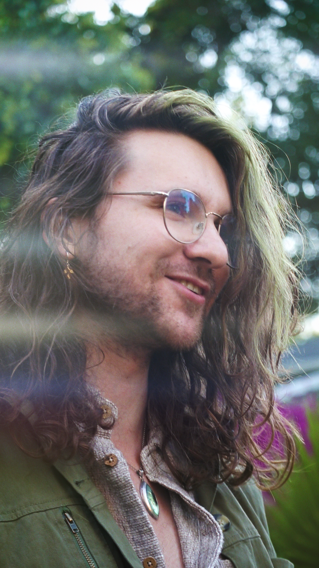 A man with long, curly hair, glasses, and a smile, outdoors with blurred trees and flowers in the background.