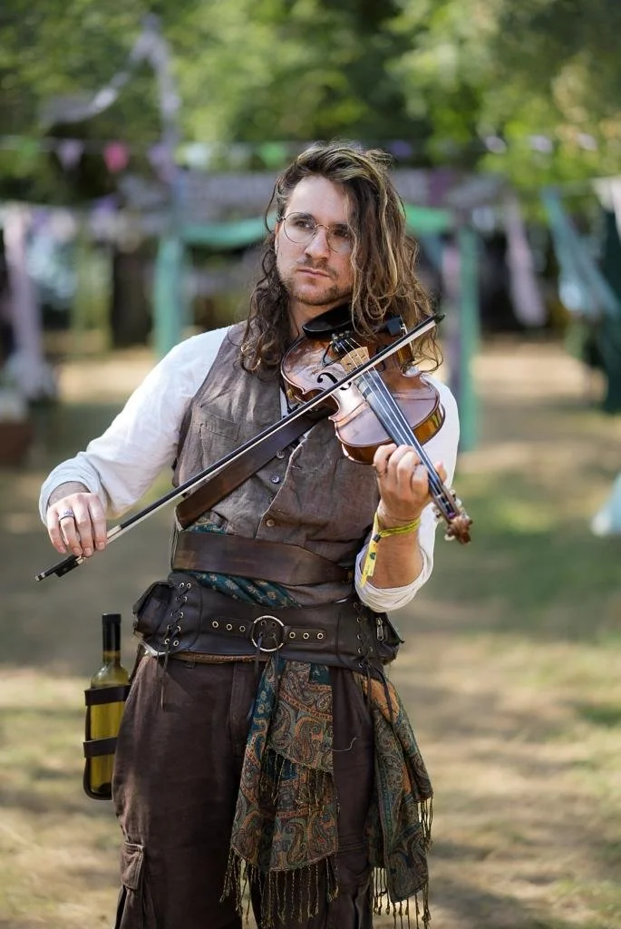 A man with long curly hair, glasses, and a vest playing a violin outdoors at a festival or gathering.