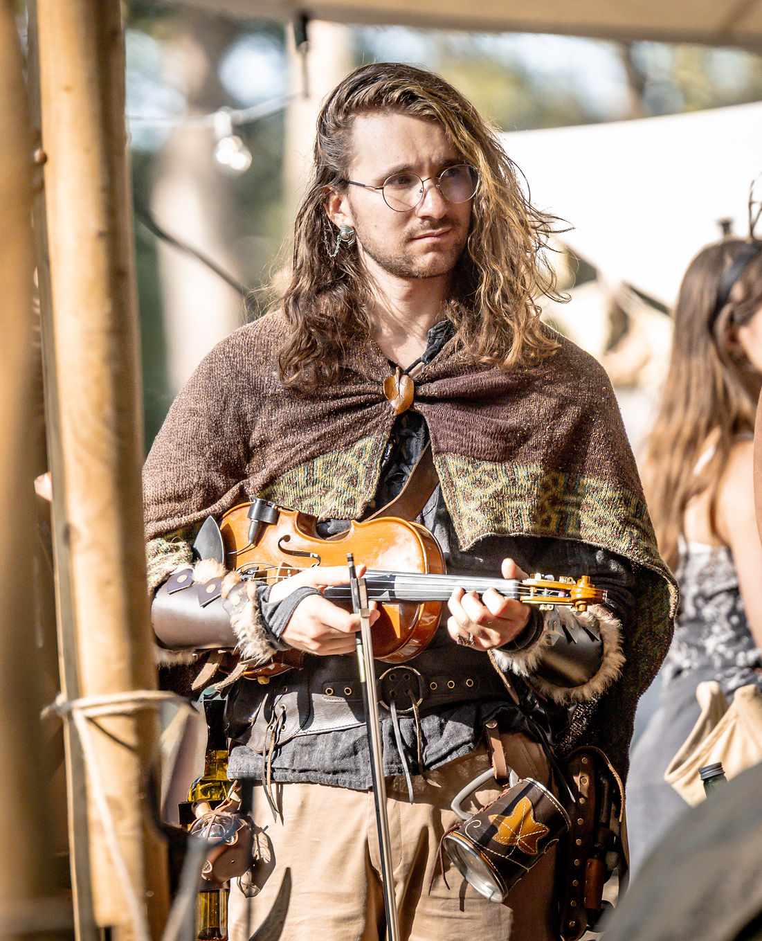 A man with long wavy hair, glasses, and earrings, dressed in medieval or fantasy-style clothing, holding a violin, on a set or outdoor location.