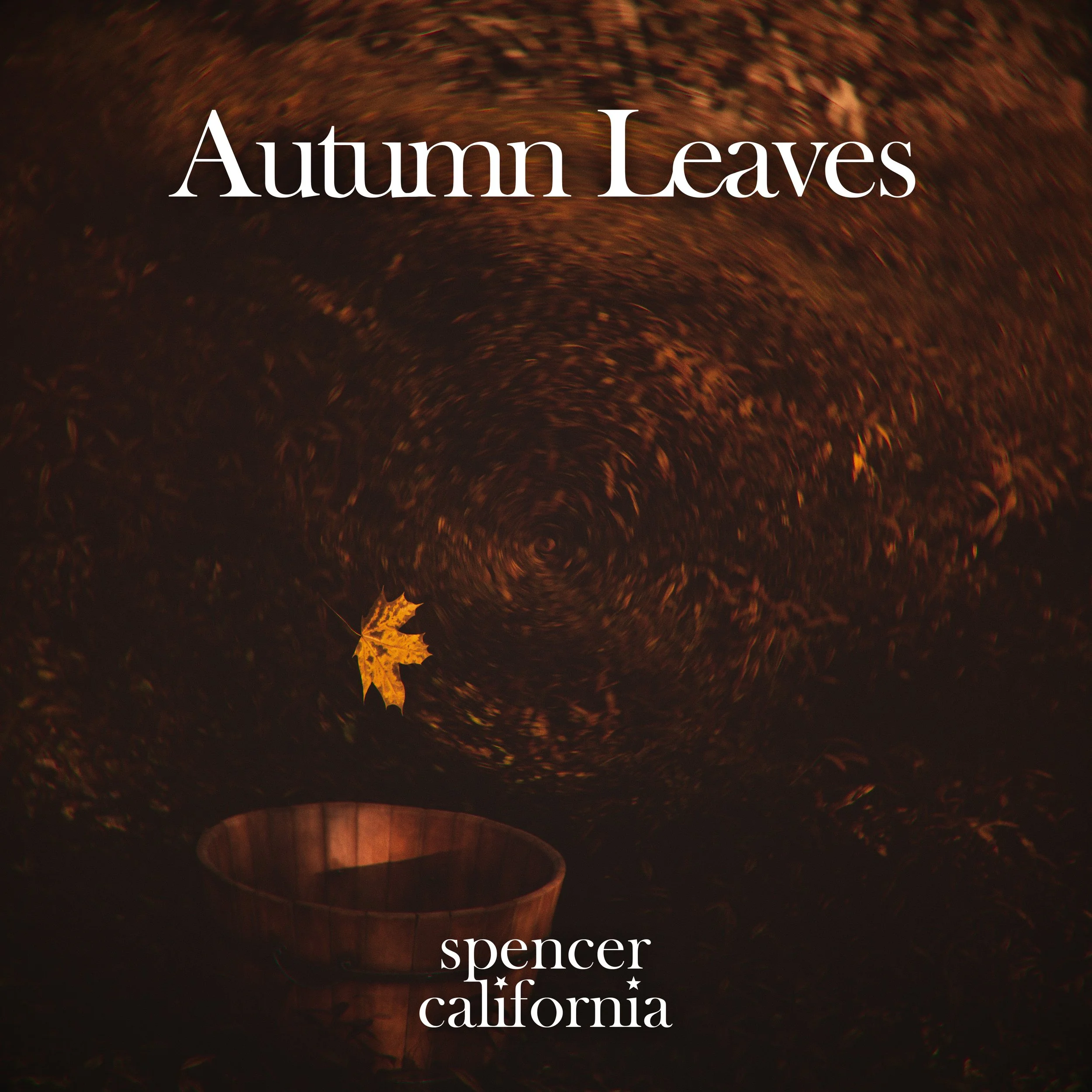 A swirling whirlpool on a water surface with fallen autumn leaves and a wooden bucket nearby. The text 'Autumn Leaves' and 'Spencer California' is overlaid on the image.