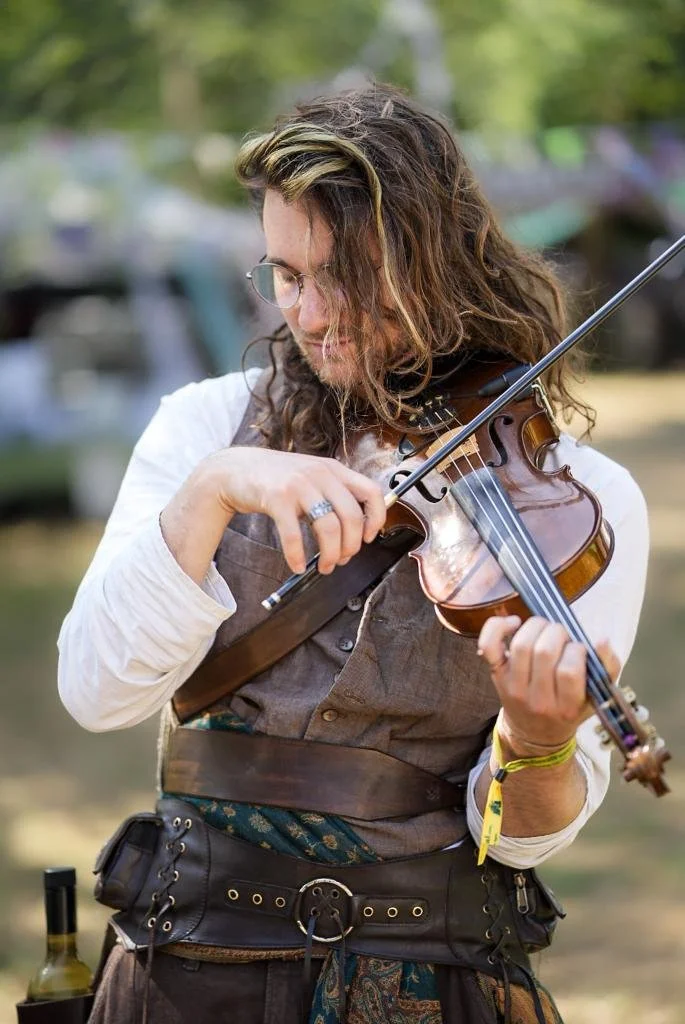 A musician playing a violin outdoors, wearing glasses, a brown vest, and a tool belt, with trees in the background.