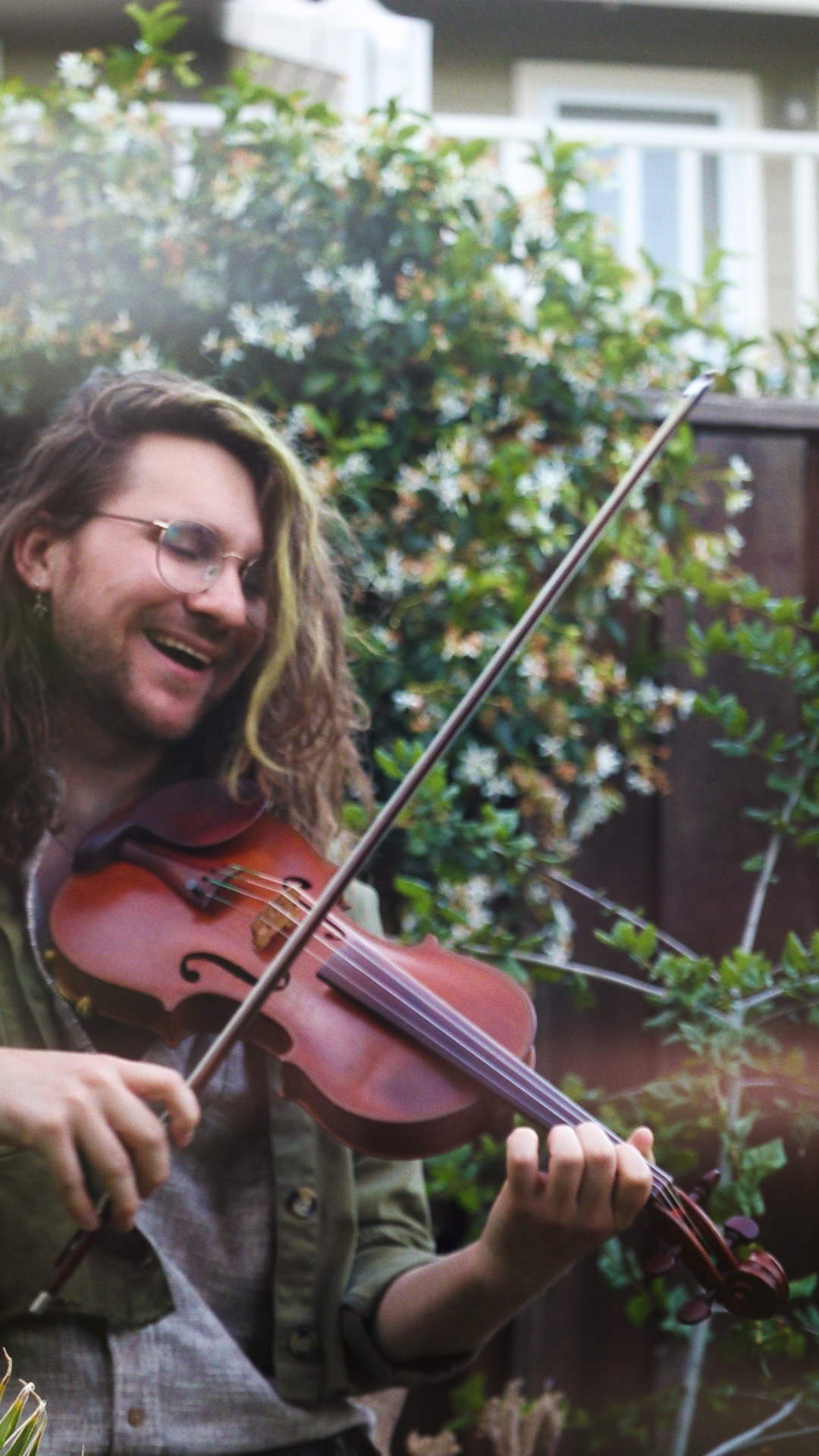 Person with long curly hair and glasses playing a violin outdoors, smiling, with greenery and flowers in the background.