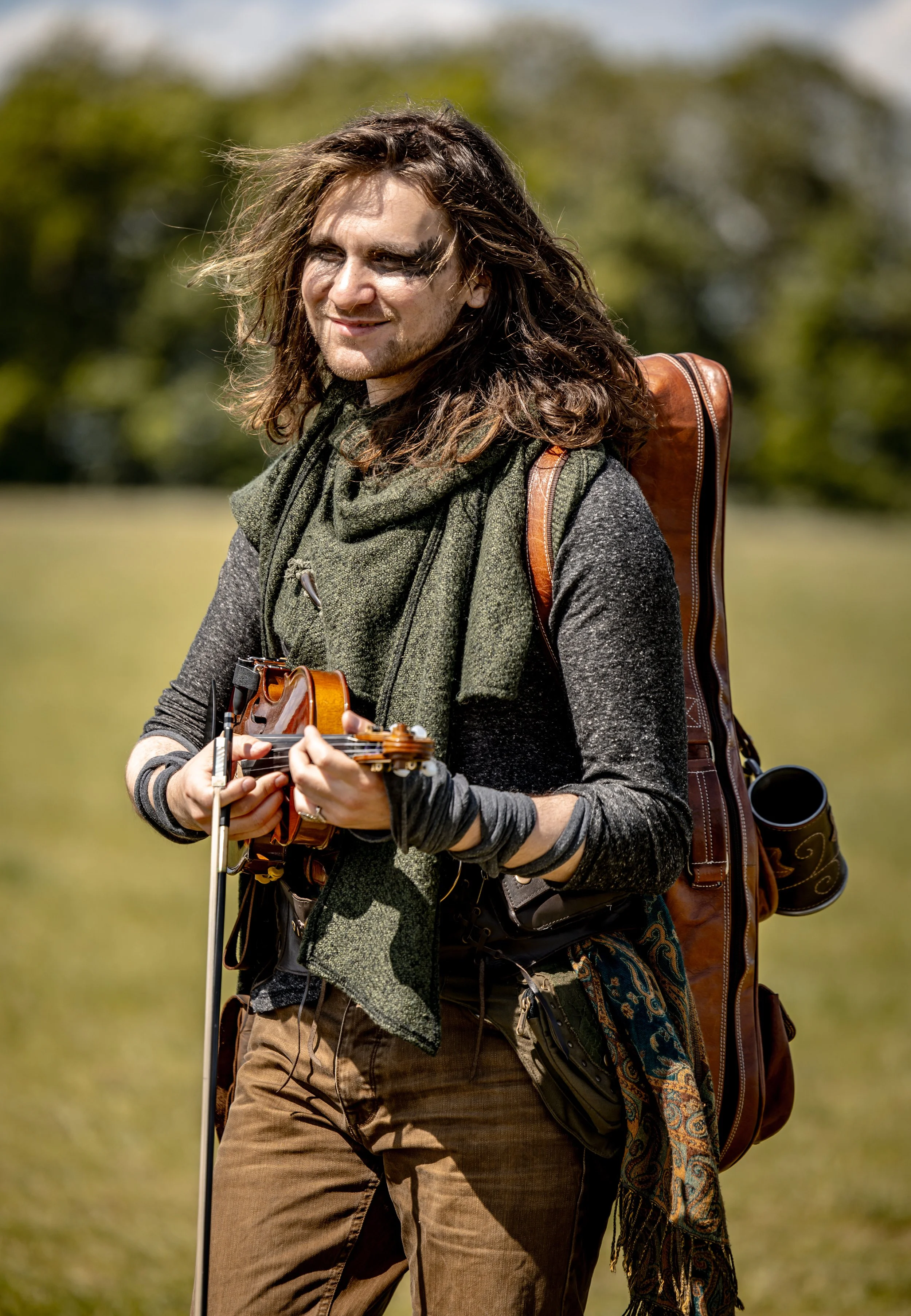 A person with long hair holding a violin outdoors in a grassy area, wearing a scarf, backpack, and casual clothing.