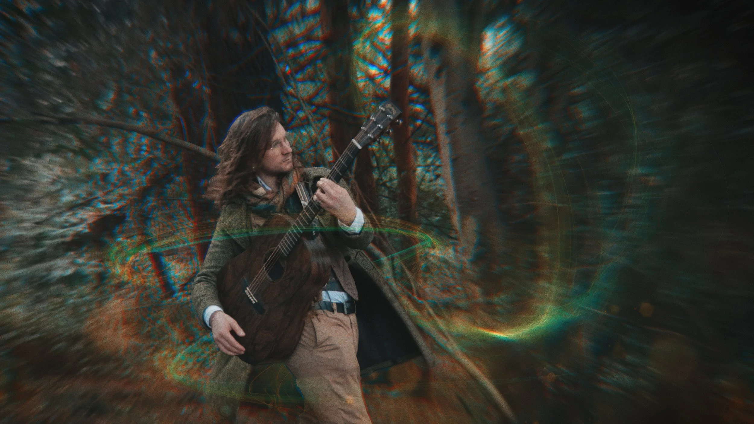 A man with shoulder-length hair and glasses walking through a blurry, abstract forest scene, playing an acoustic guitar.