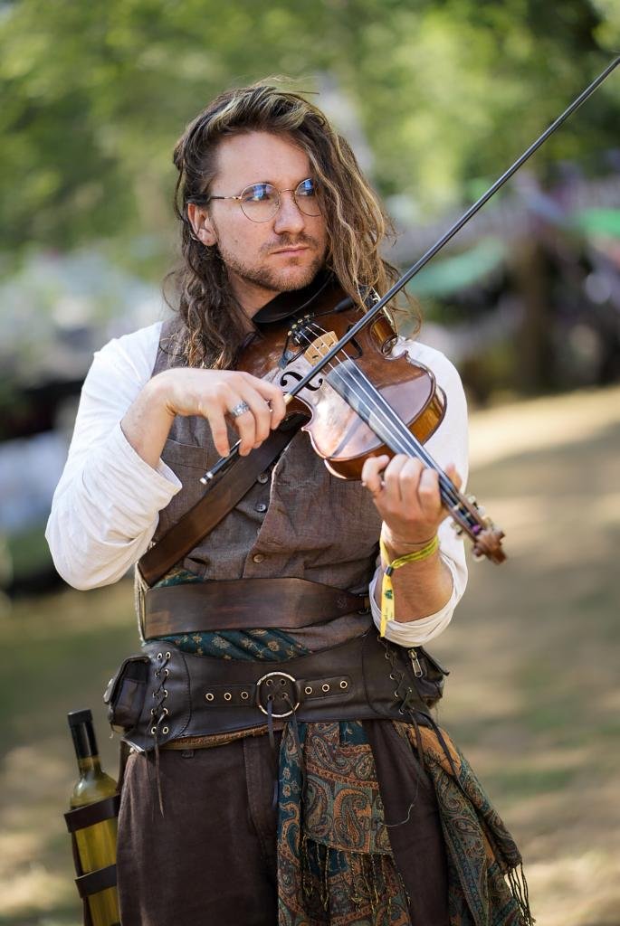 A man with long curly hair, glasses, and a beard, playing a violin outdoors.
