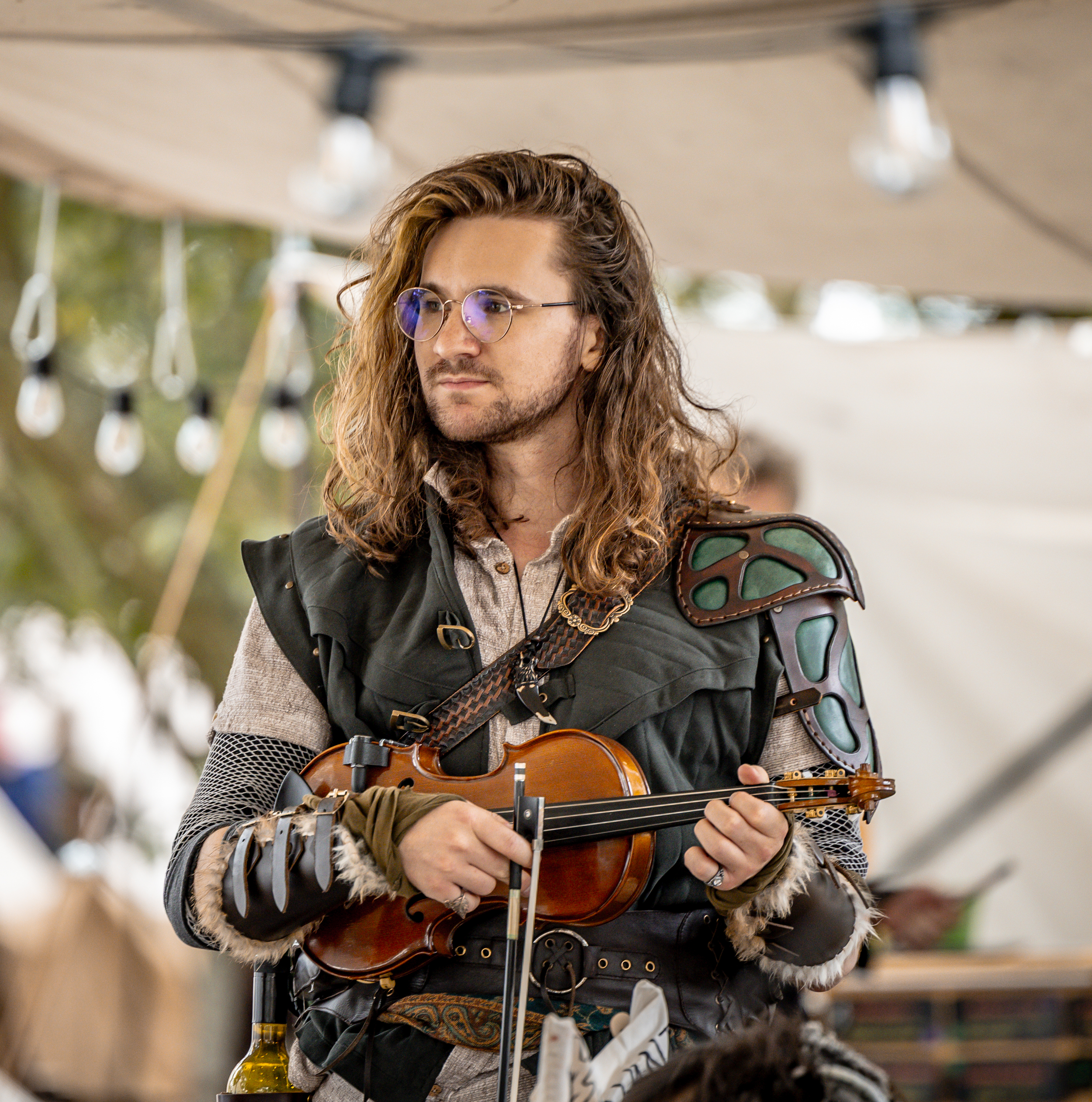 A man with long, curly hair and glasses plays a violin at an outdoor event, wearing medieval or fantasy-style armor and accessories.