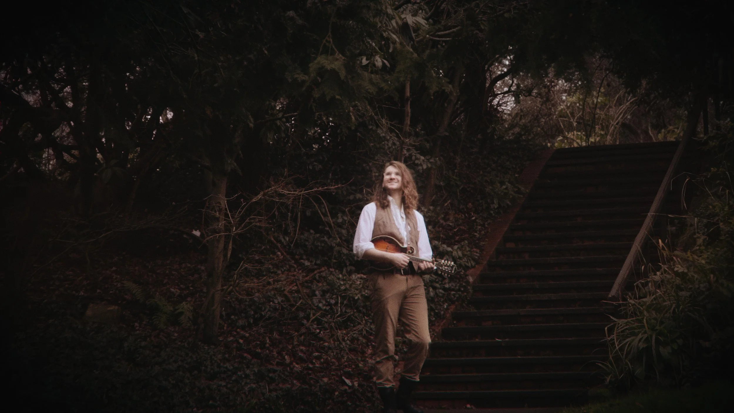 A young man with long, curly hair standing outdoors at dusk or dawn, holding a mandolin, smiling, surrounded by trees, near a staircase with dark steps and a metal railing.