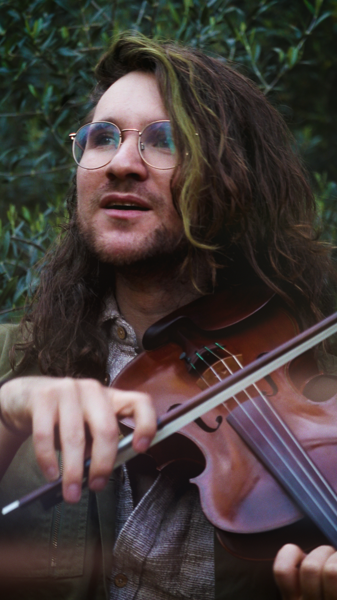 A person with long, curly hair and glasses playing a violin outdoors with a background of green foliage.
