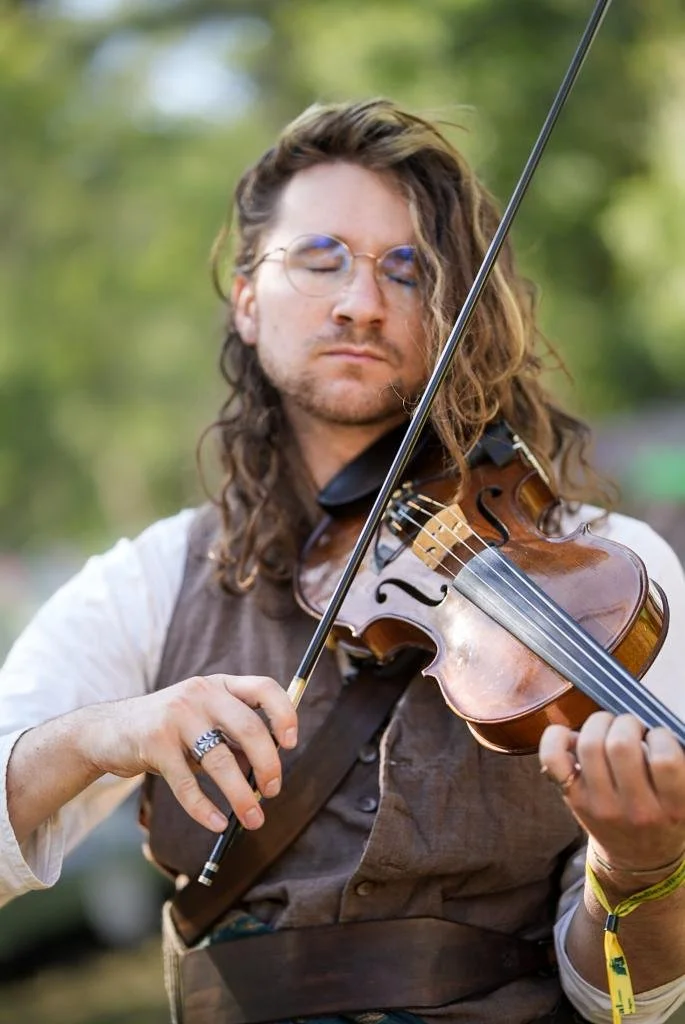 A man with glasses and long, wavy hair playing a violin outdoors.