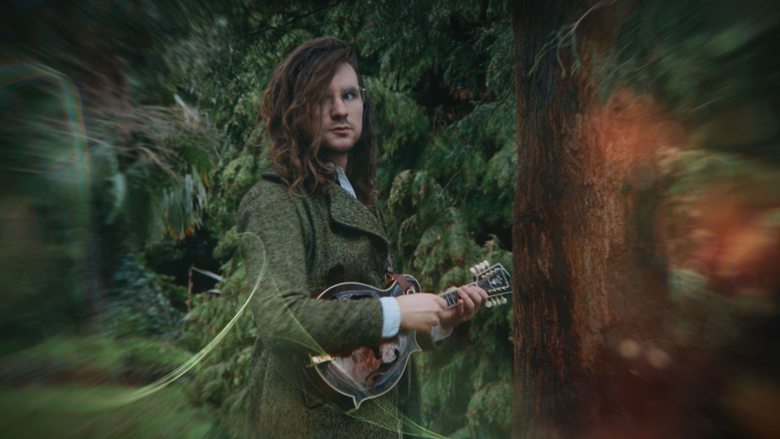 Man with long hair playing mandolin in a forest.
