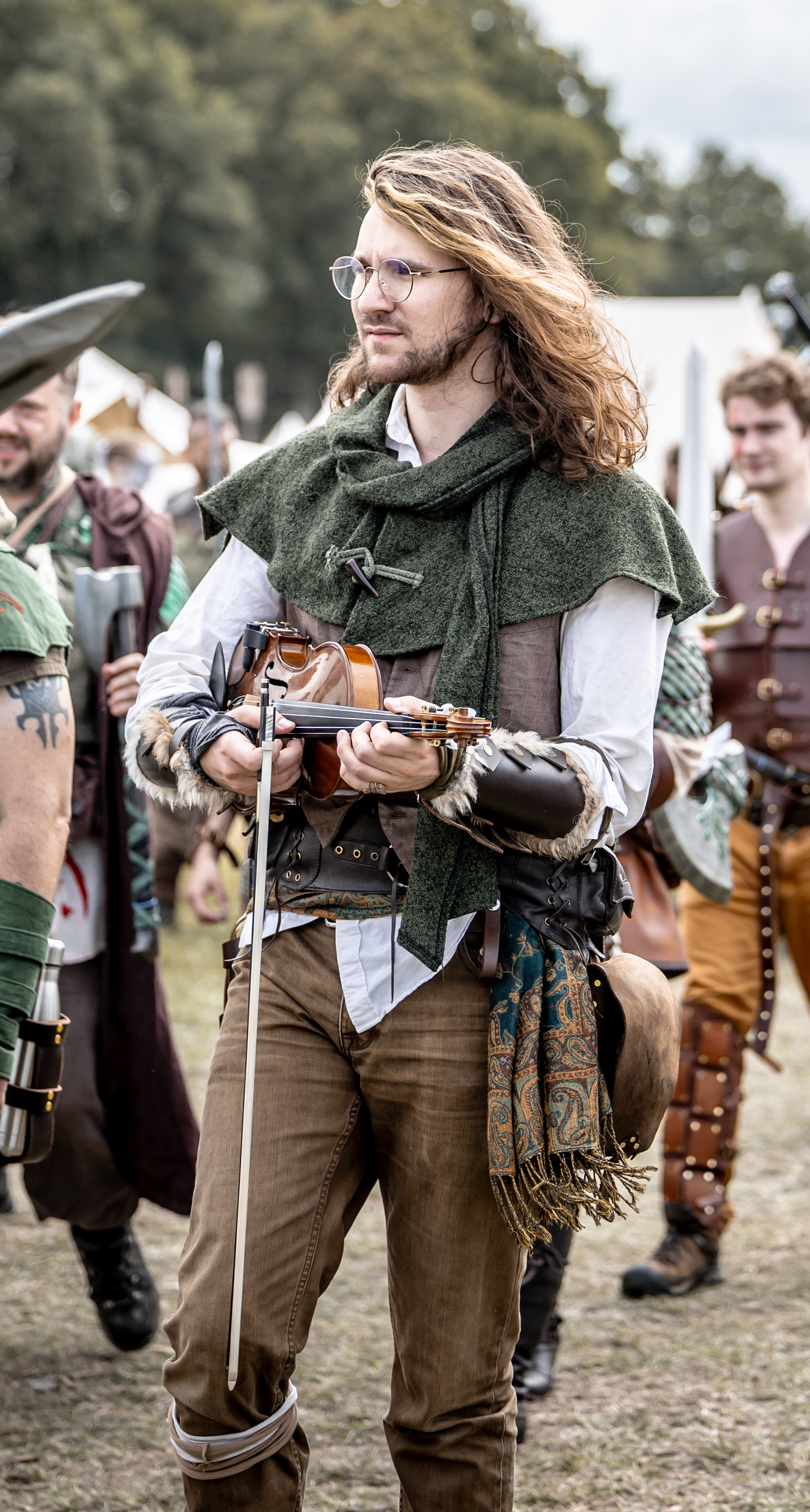 A man with long hair and glasses holding a violin and bow, dressed in medieval-style clothing, with others in similar costumes in the background at a historical reenactment event.