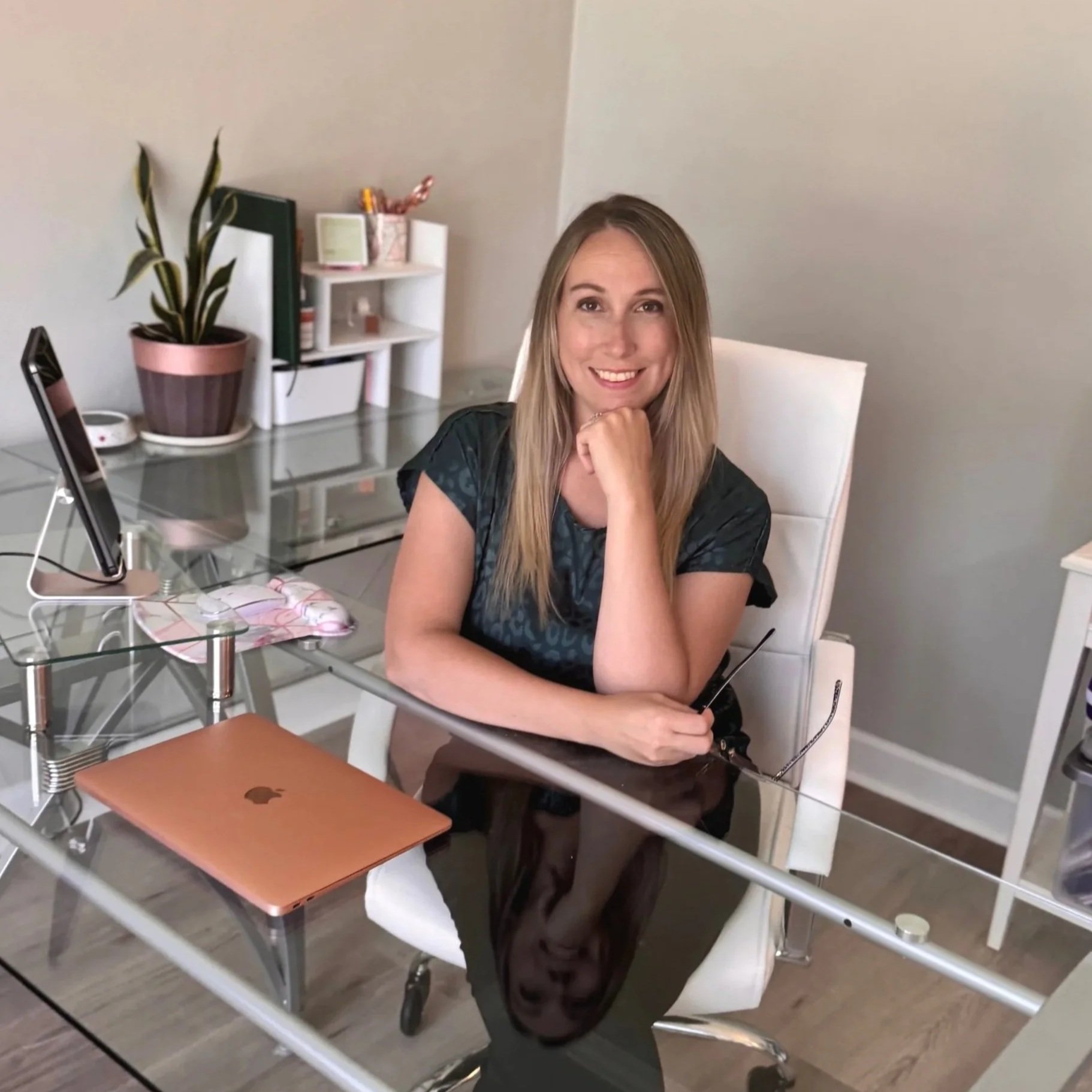 Woman sitting at a glass desk with a closed rose gold laptop, holding glasses, smiling, in a home office with a plant, books, and a white chair.