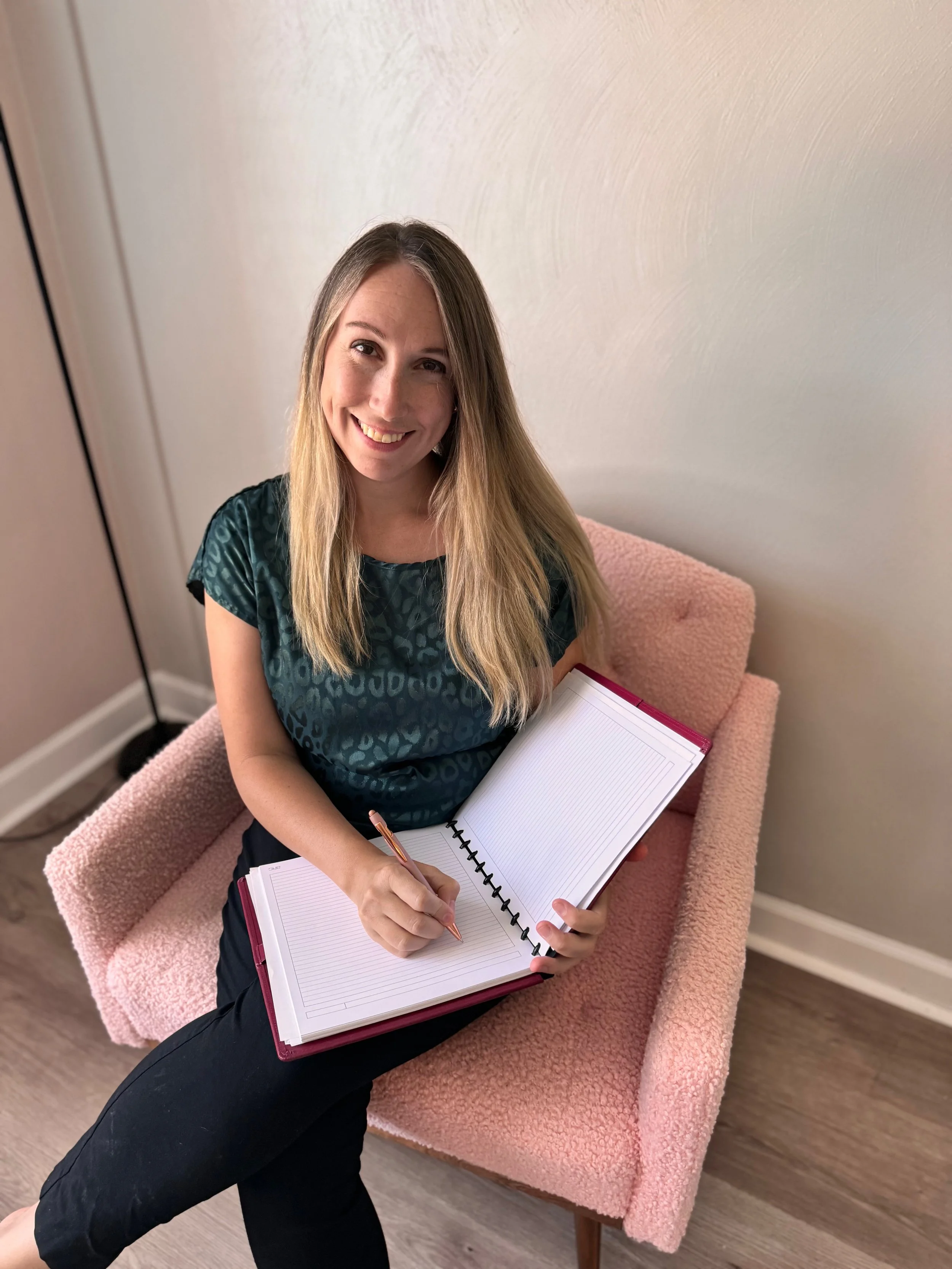 A woman with long blonde hair sitting on a pink textured armchair, smiling at the camera, holding an open pink notebook with a spiral binding, and writing with a pen.