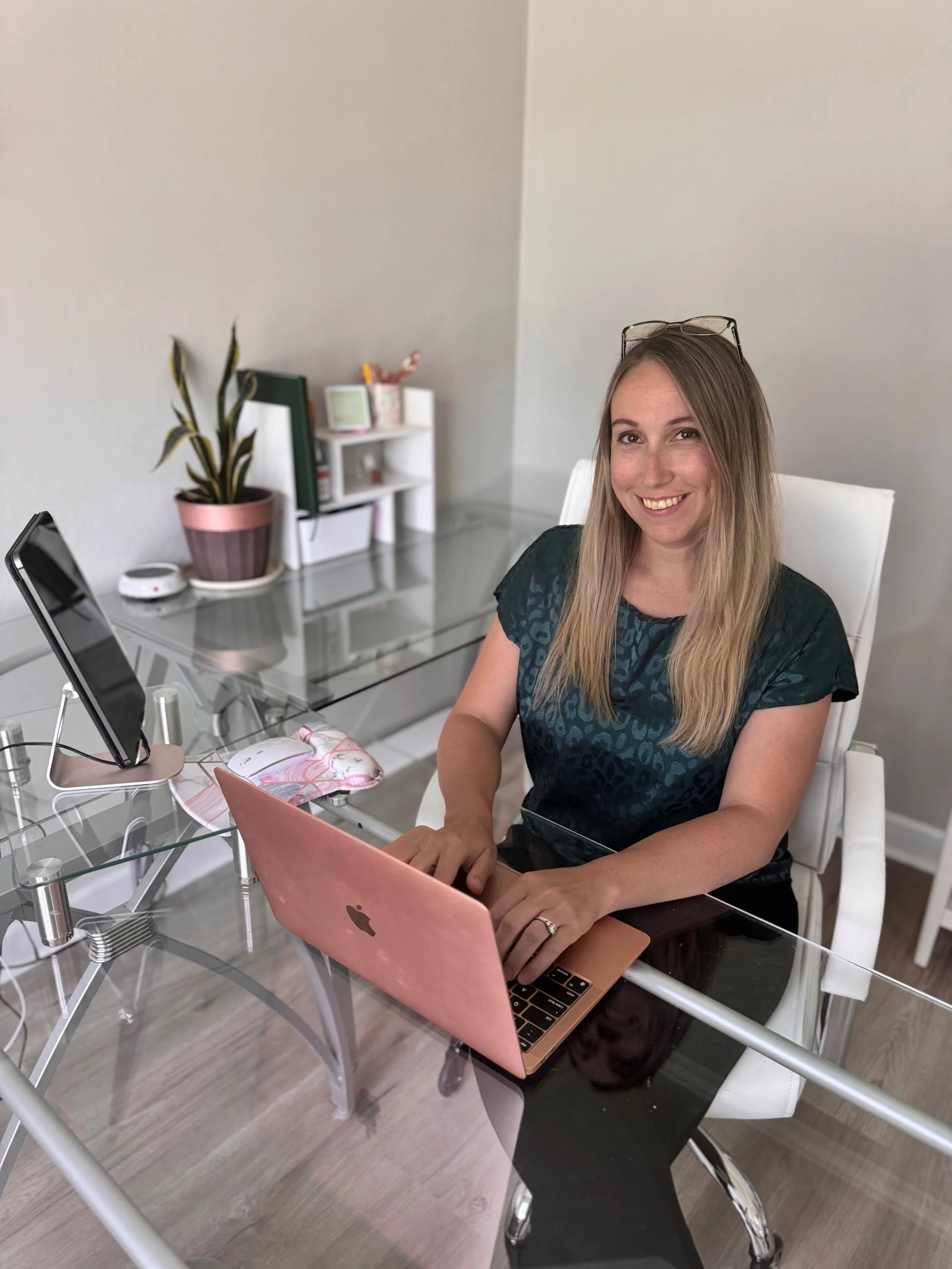 A woman with long blonde hair wearing a dark green patterned shirt sits at a glass desk using a rose gold MacBook. She is smiling and has reading glasses on her head. On the desk are a pink mouse pad with a mouse, a monitor on a stand, and a smartpho