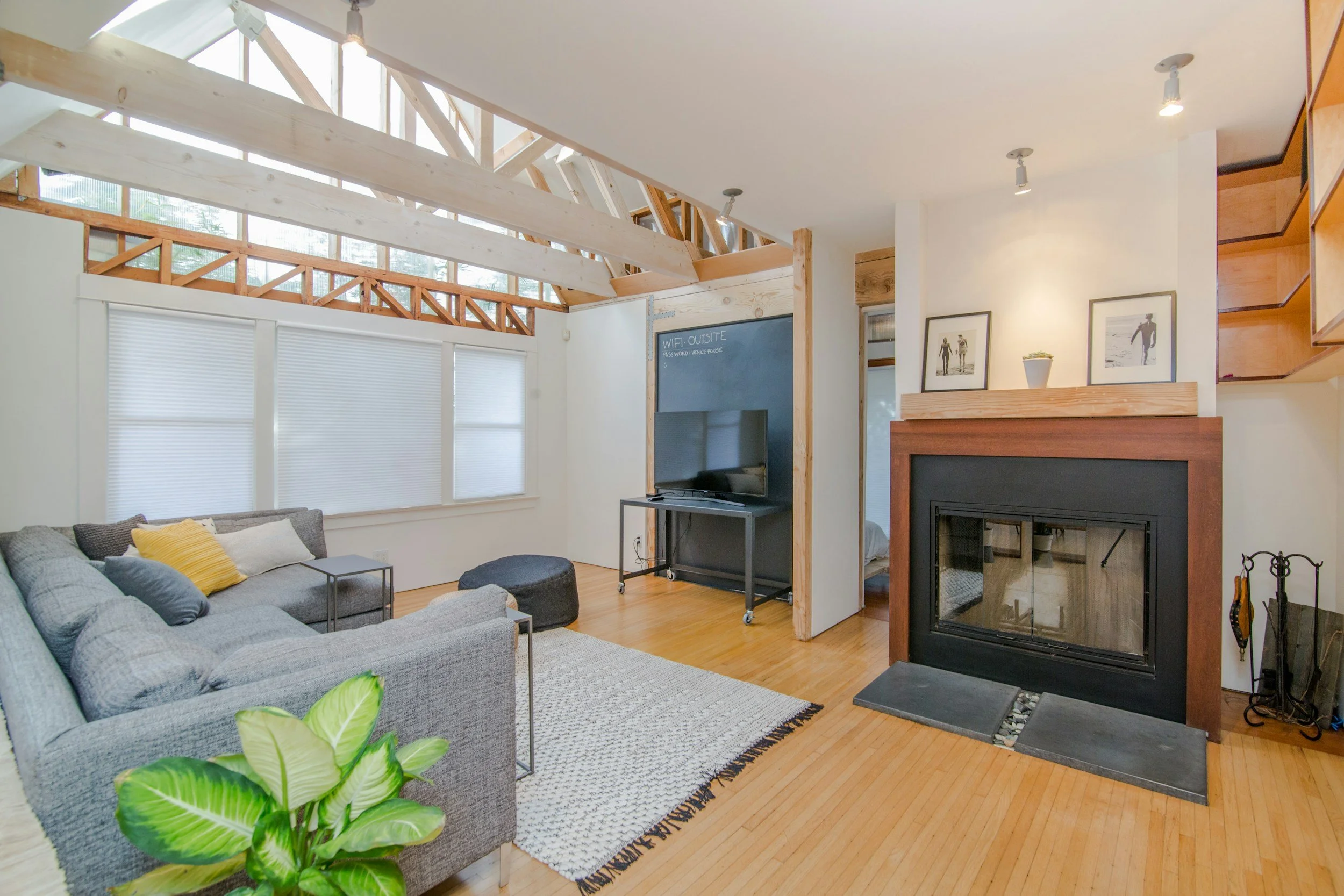 Living room with a gray sectional sofa, a black ottoman, a television on a stand, and a fireplace with wood and glass doors. The room has high ceilings with exposed wooden beams and large windows with white blinds.