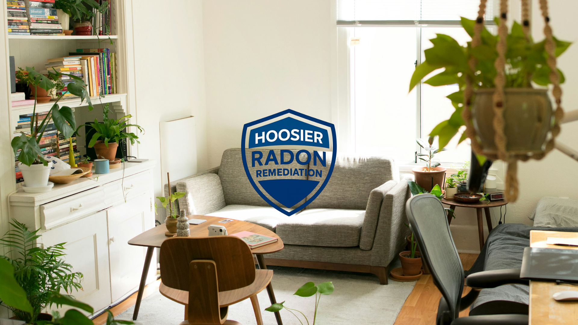 Living room with a gray sofa, wooden table, bookshelves, and various houseplants. A logo with the text 'Hoosier Radon Remediation' is centered in the image.