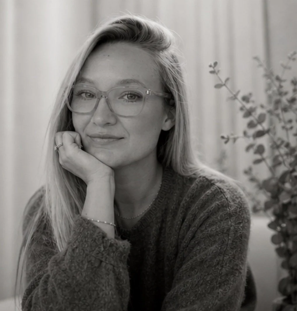 A black-and-white portrait of Sherri LaRocque with glasses, resting her chin on her hand and smiling slightly, with a plant in the background.