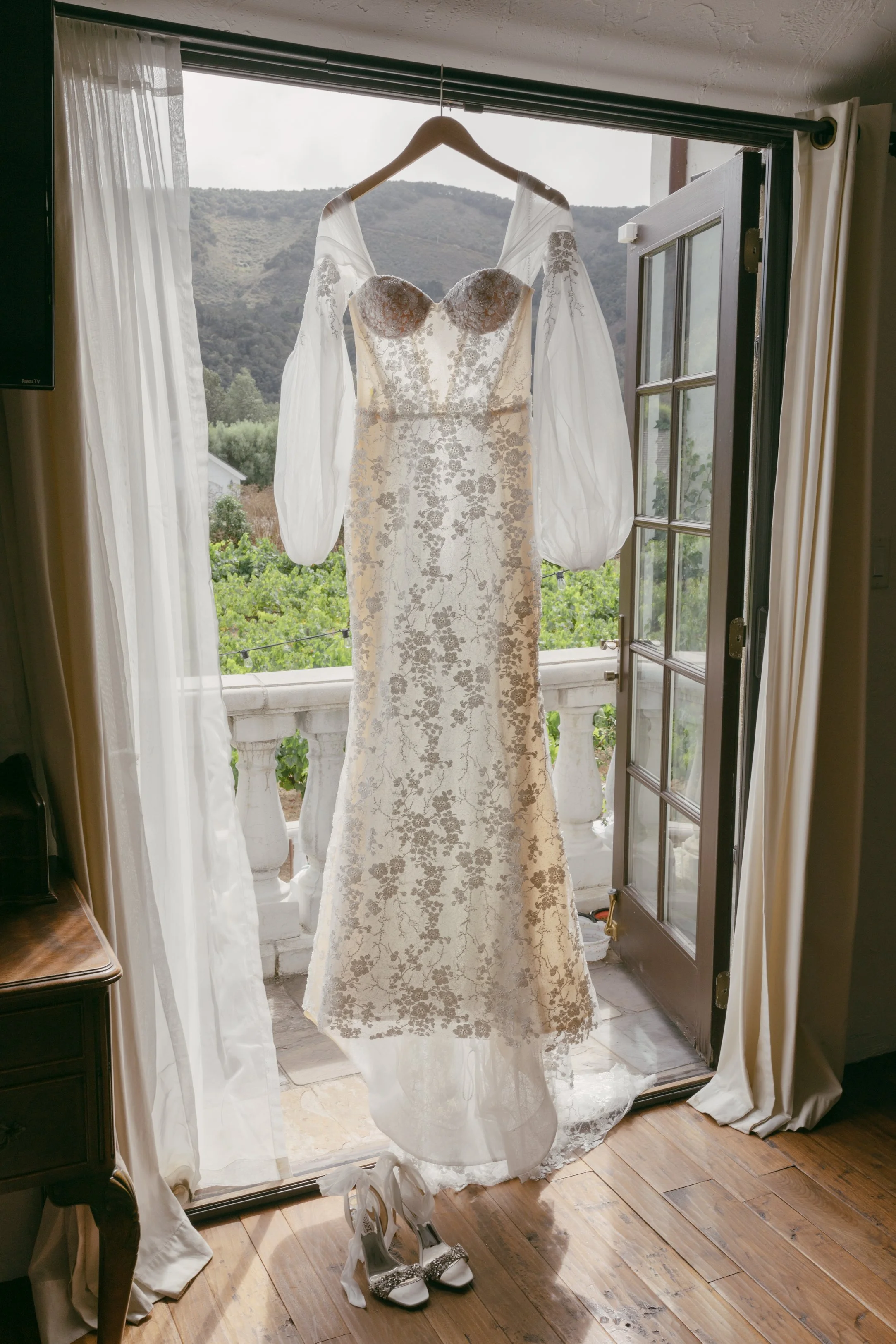Wedding dress hanging in front of open glass door, with high heels placed underneath, overlooking a scenic mountain view.