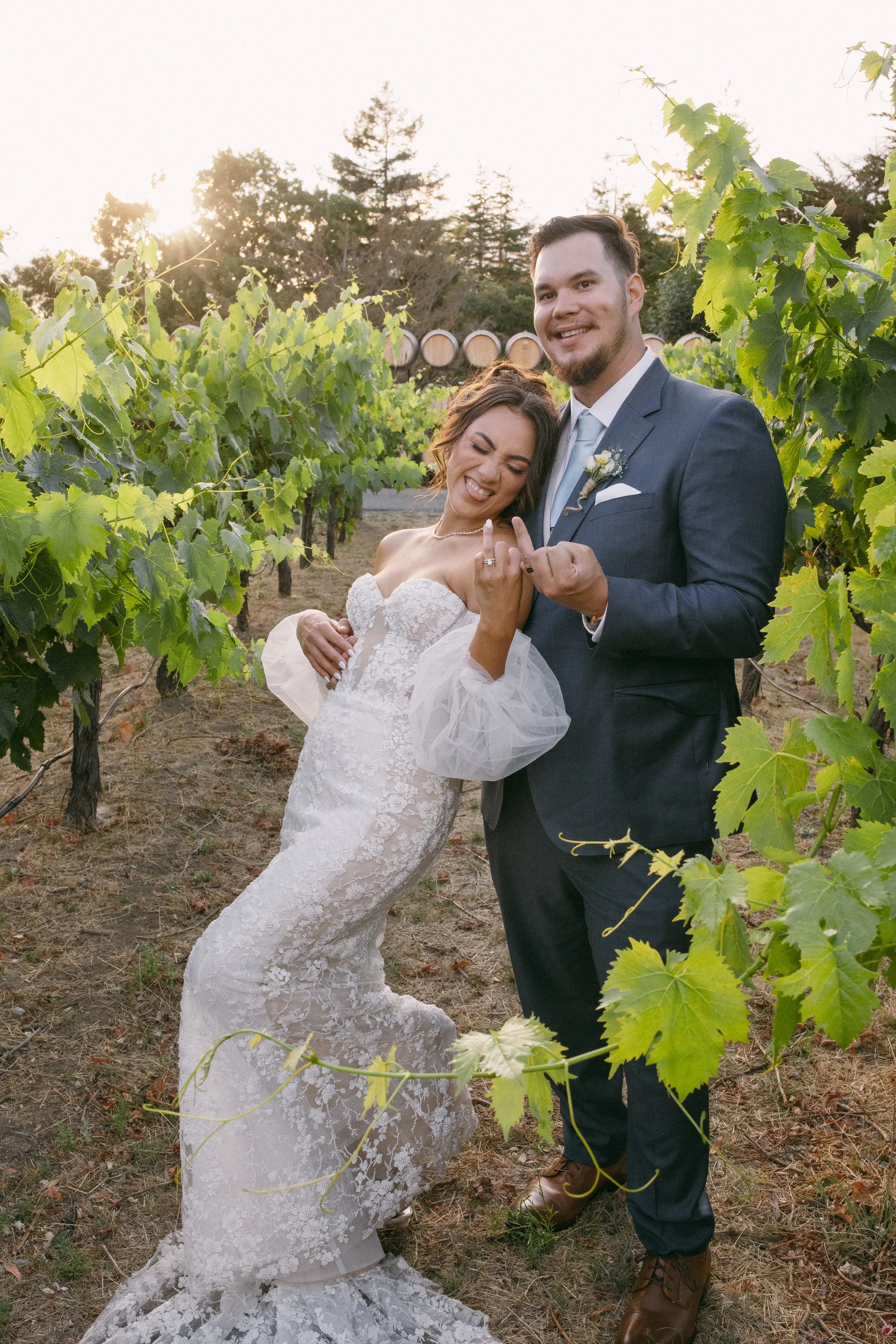 A newlywed couple in wedding attire pose in a vineyard, showing their wedding rings, with the bride smiling and the groom smiling, the setting sun in the background.