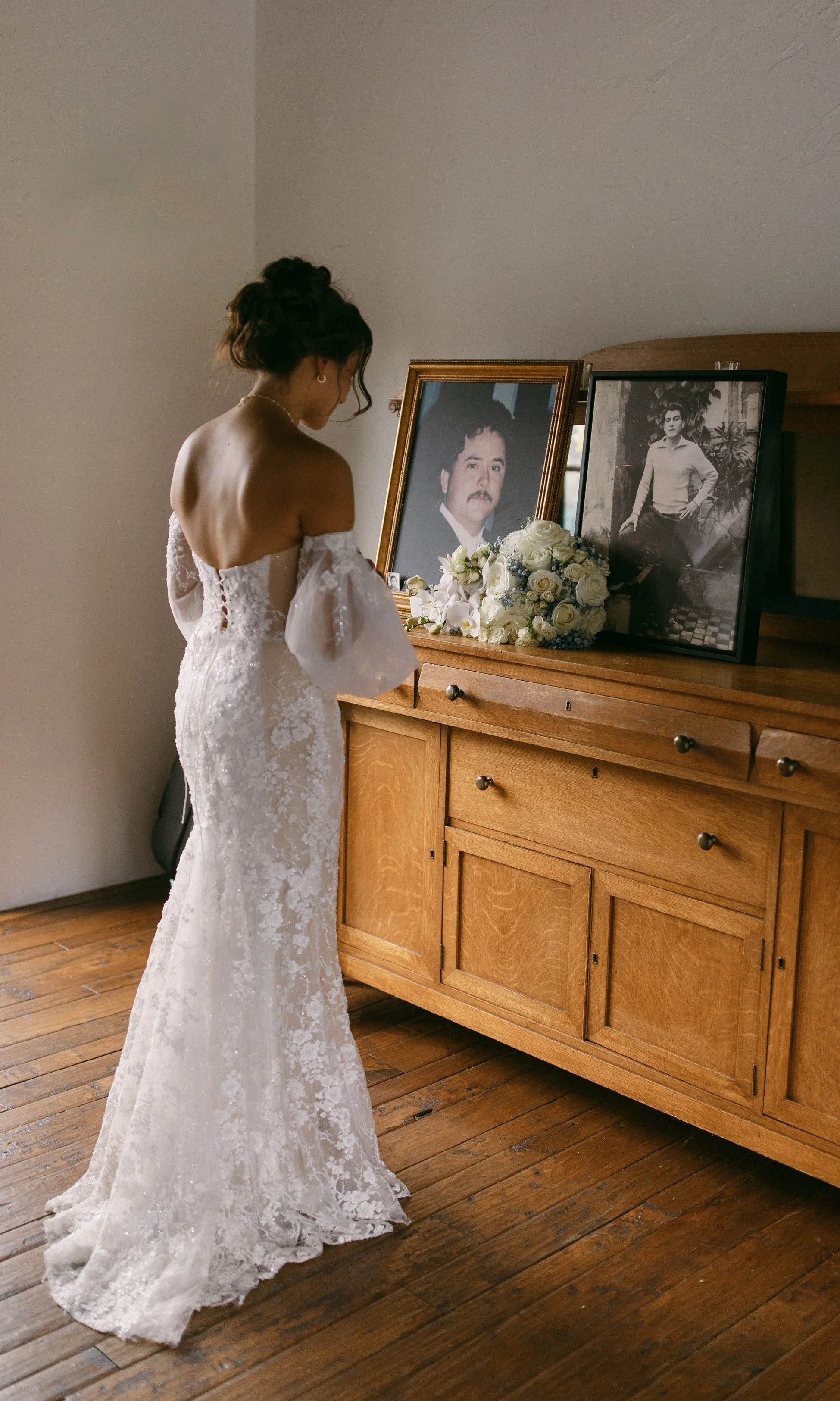 A woman in a white wedding dress standing in front of a wooden cabinet with framed photographs and a bouquet of white roses, seemingly in a moment of reflection or prayer.