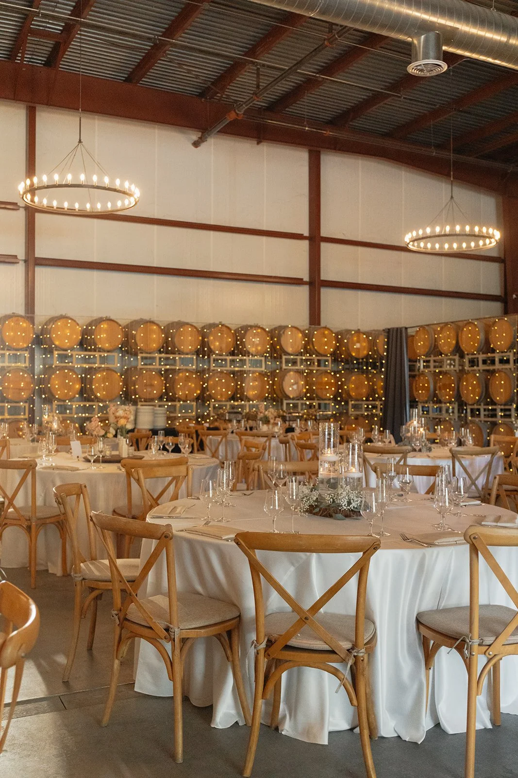 Interior of a banquet hall with round tables covered in white tablecloths, set with glassware and floral centerpieces, surrounded by wooden chairs, and decorated with wine barrel wall accents and string lights.