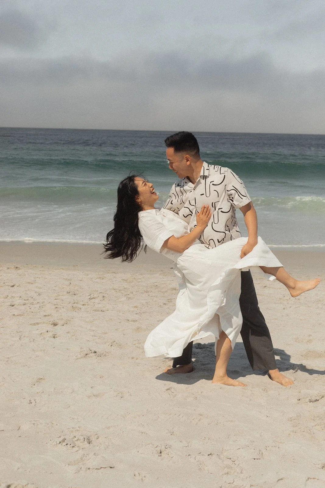 A man lifting a woman on a beach, both smiling and enjoying each other's company, with the ocean in the background.
