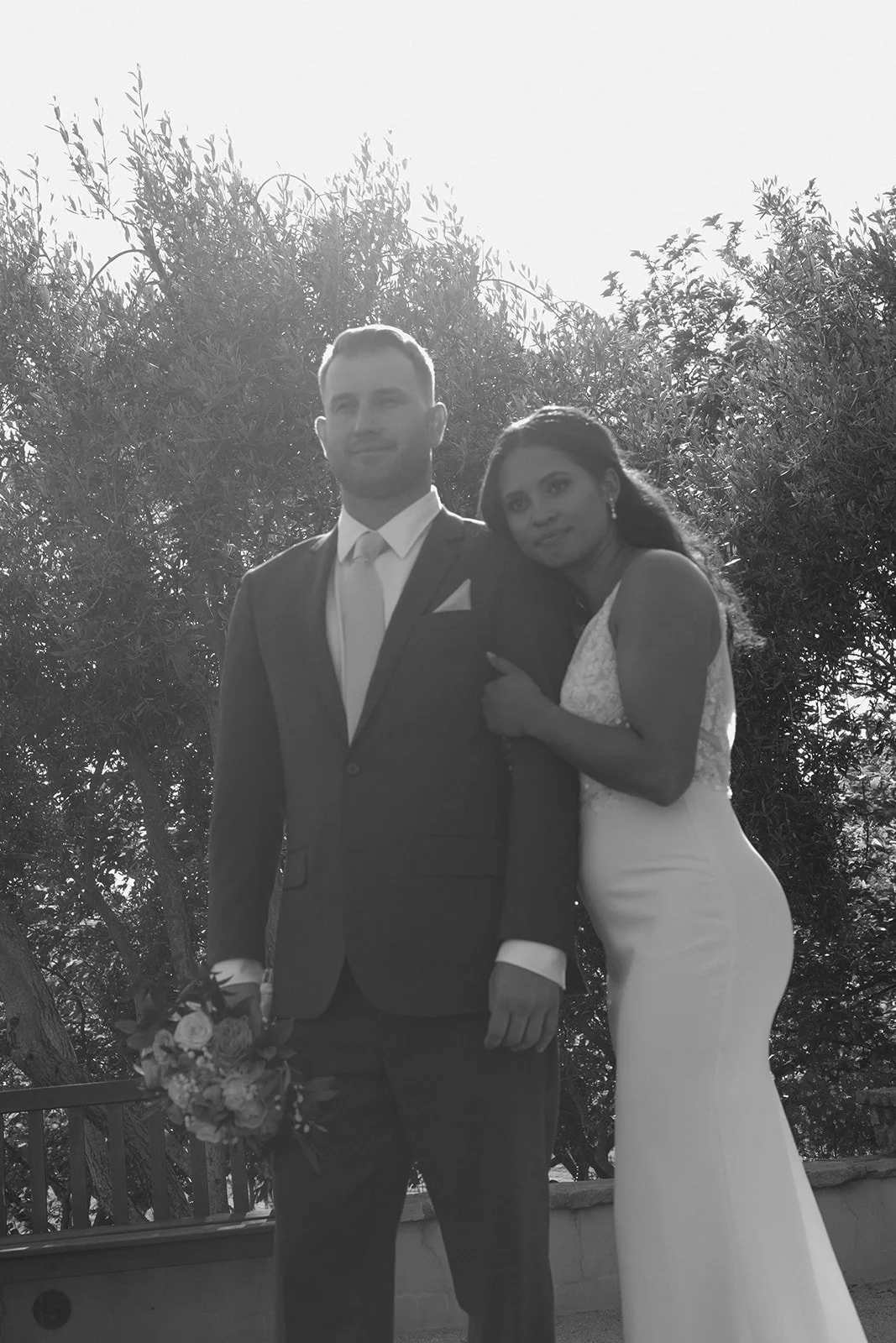 Black and white photo of a newlywed couple outdoors, the man in a suit holding a bouquet of flowers, and the woman in a white wedding dress leaning on his shoulder, with trees in the background.