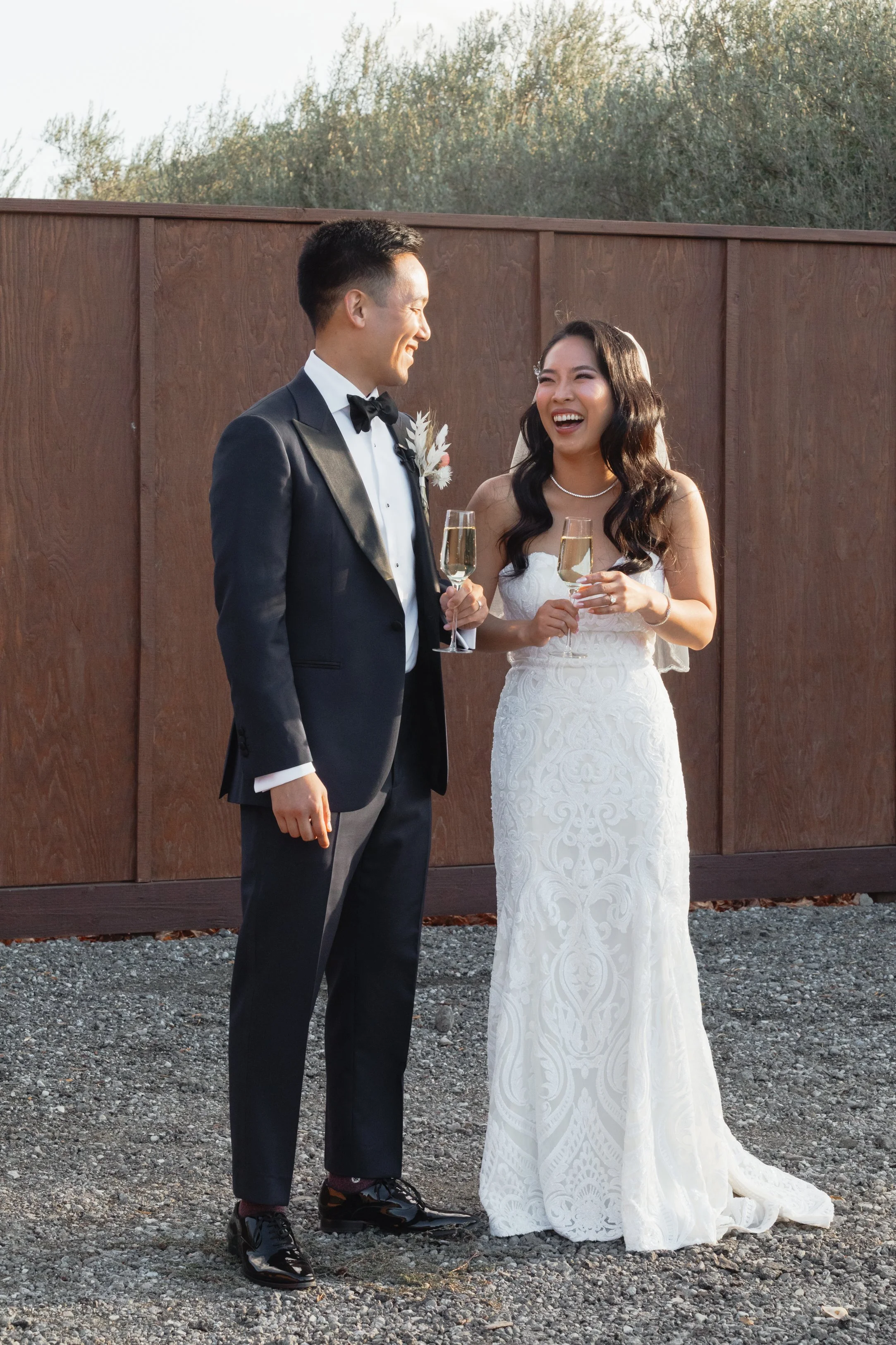 A bride and groom in wedding attire sharing a laugh while holding glasses of champagne outdoors.