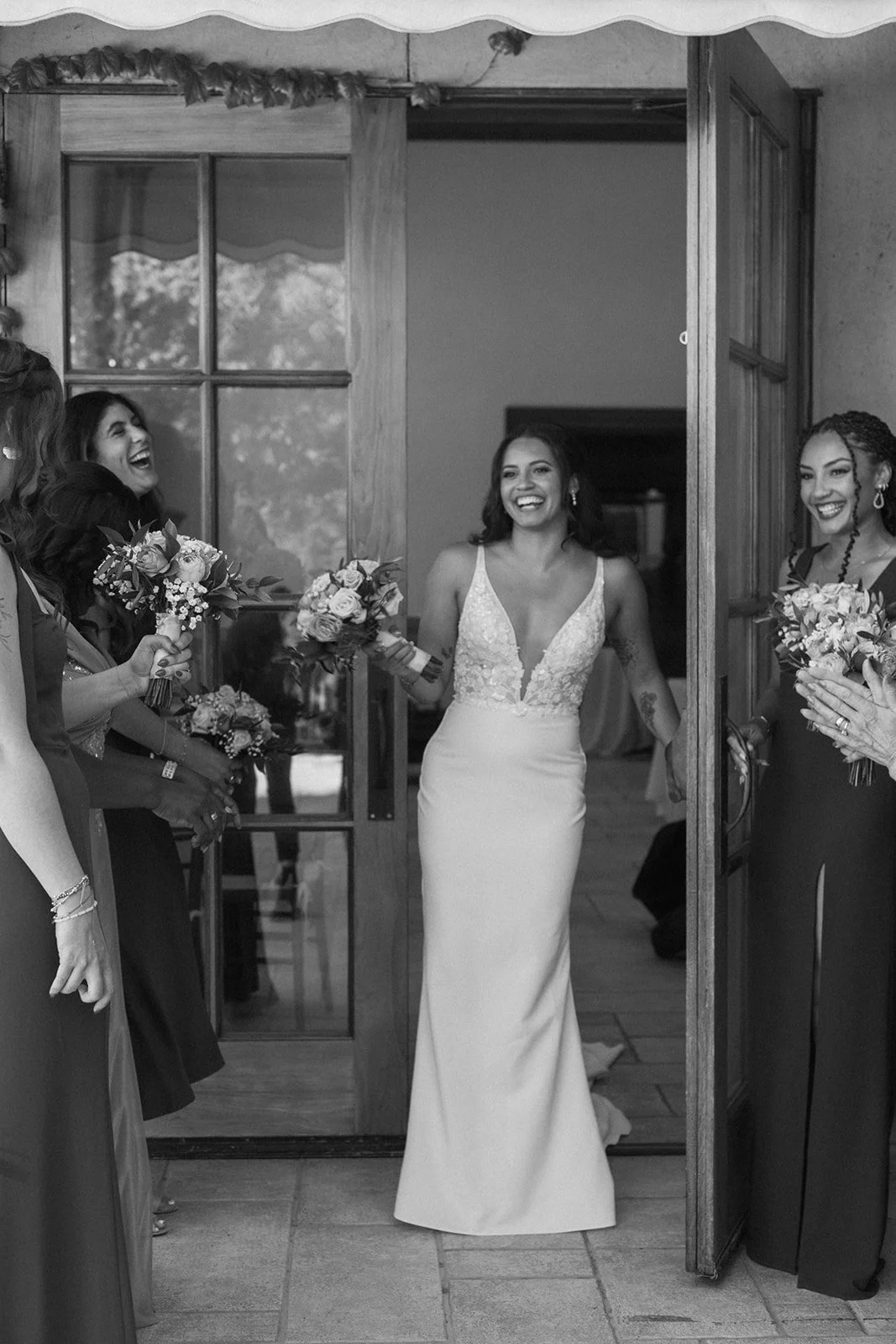 A bride in a wedding dress walking into a room while her bridesmaids and friends smile and hold bouquets.